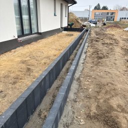 P.H.U Konrad Pacholczyk Metrycki - Wkopane obrzeża z szarej kostki betonowej oddzielające teren pod trawnik od rabaty, w tle budynek i pracownik z taczką. Widok z perspektywy ziemi.