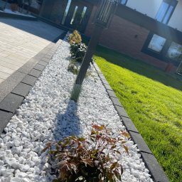 P.H.U Konrad Pacholczyk Metrycki - Ogród z białymi kamieniami, niskimi roślinami i lampą wzdłuż ścieżki. Widok na dom z trawnikiem. Elementy małej architektury ogrodowej.