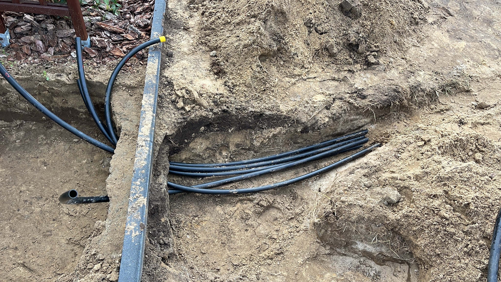 Wykop z ułożonymi czarnymi rurami, prawdopodobnie instalacja nawadniająca lub elektryczna, widoczny profil ziemi i fragment obrzeża trawnika.