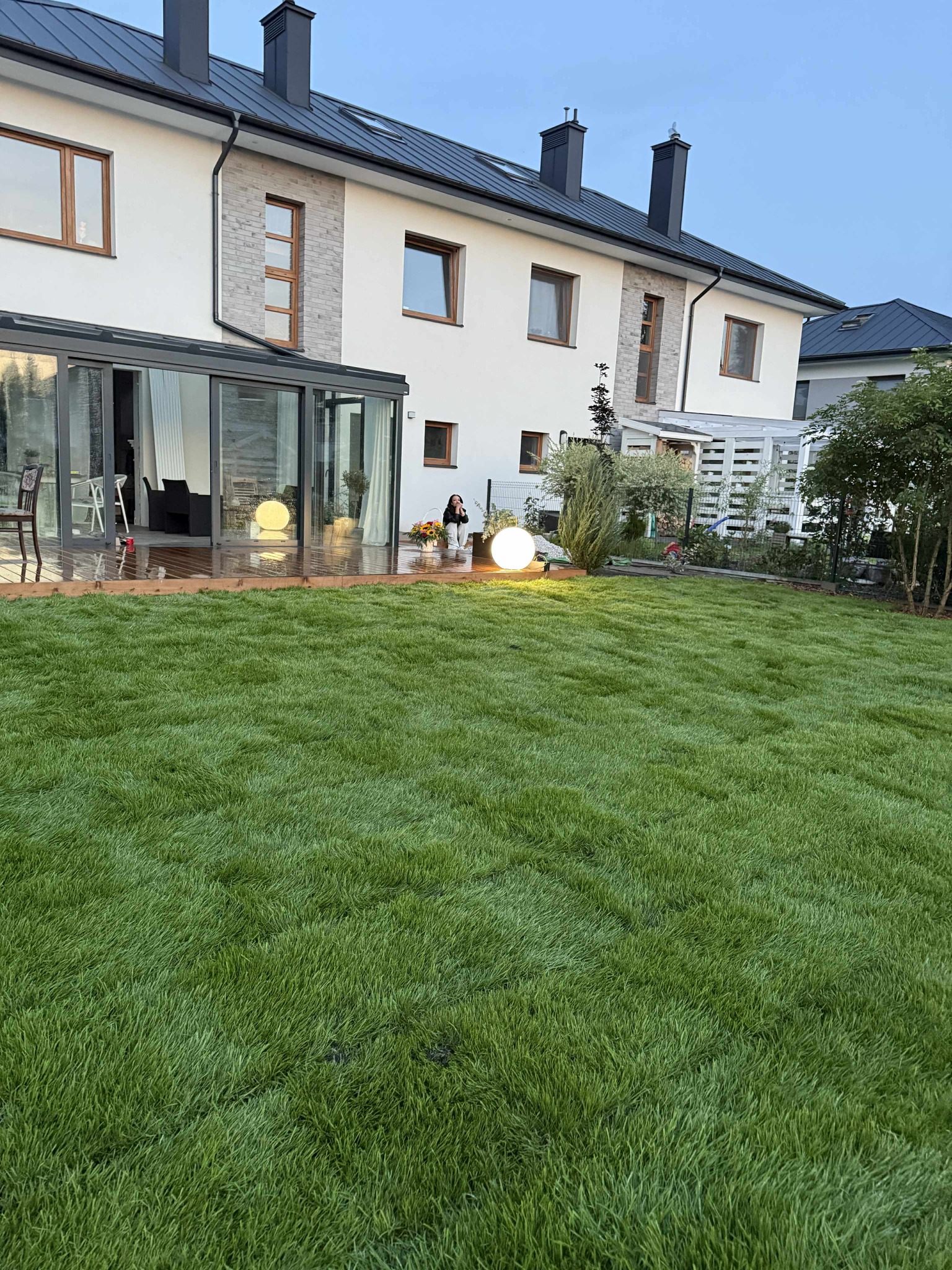 Świeżo założony trawnik z rolki przed nowoczesnym domem z przeszkloną werandą. Dekoracyjna kula świetlna i osoba z koszem kwiatów dodają uroku.