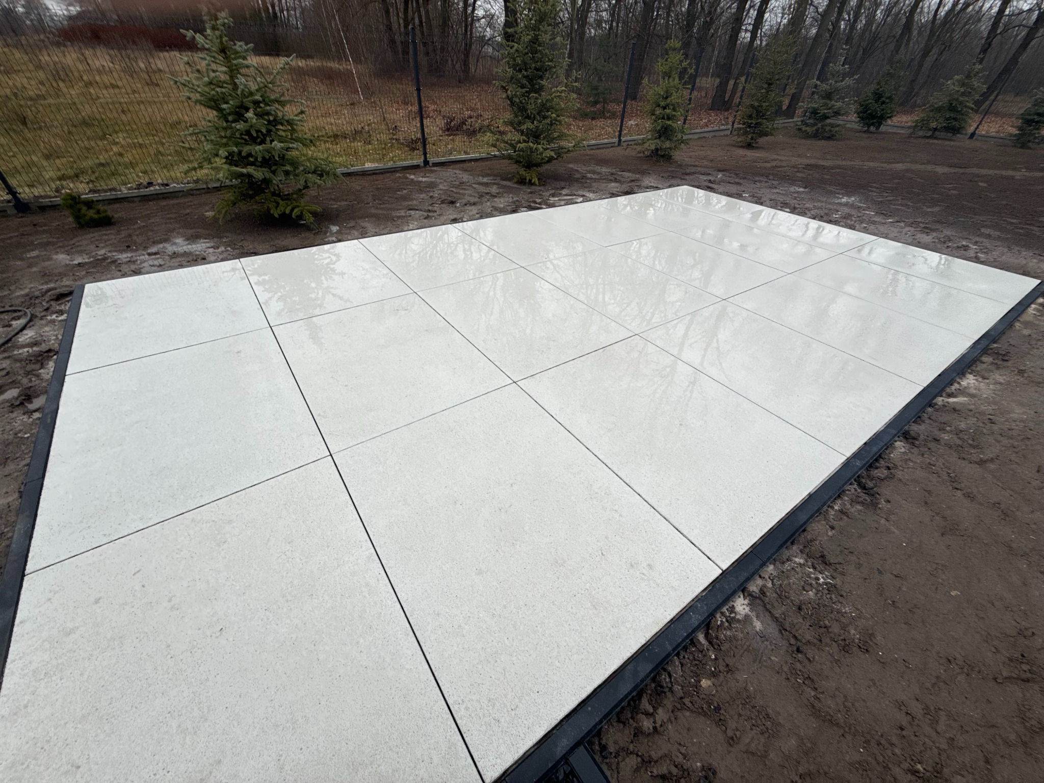 Jasne, kwadratowe płyty tarasowe ułożone na czarnej ramie, otoczone ziemią i młodymi drzewkami iglastymi. Widok z góry, pochmurna pogoda.