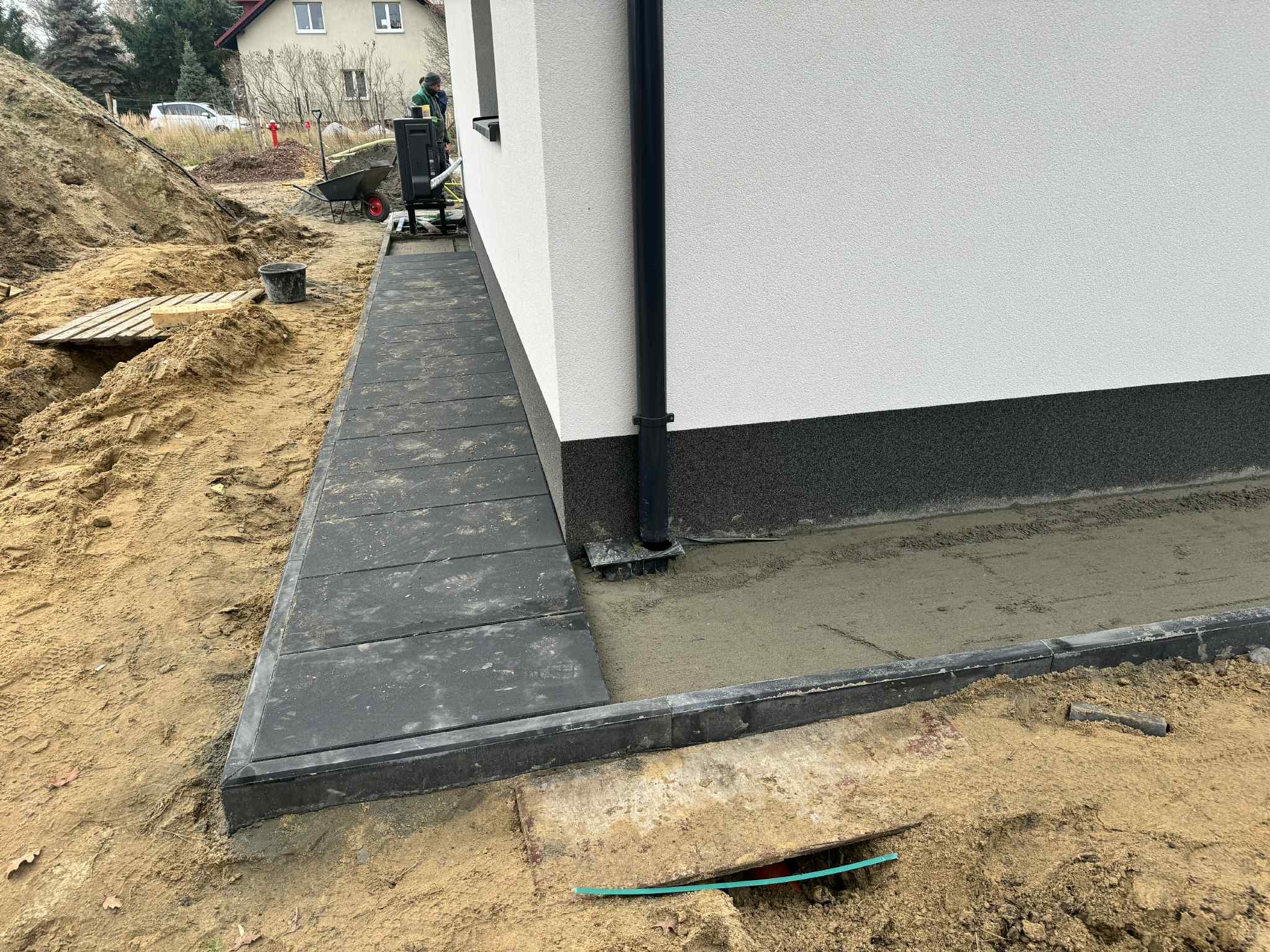 Układanie ciemnoszarej kostki brukowej wzdłuż białej ściany budynku. Widoczny fragment ogrodu w trakcie prac ziemnych i przygotowania podłoża.