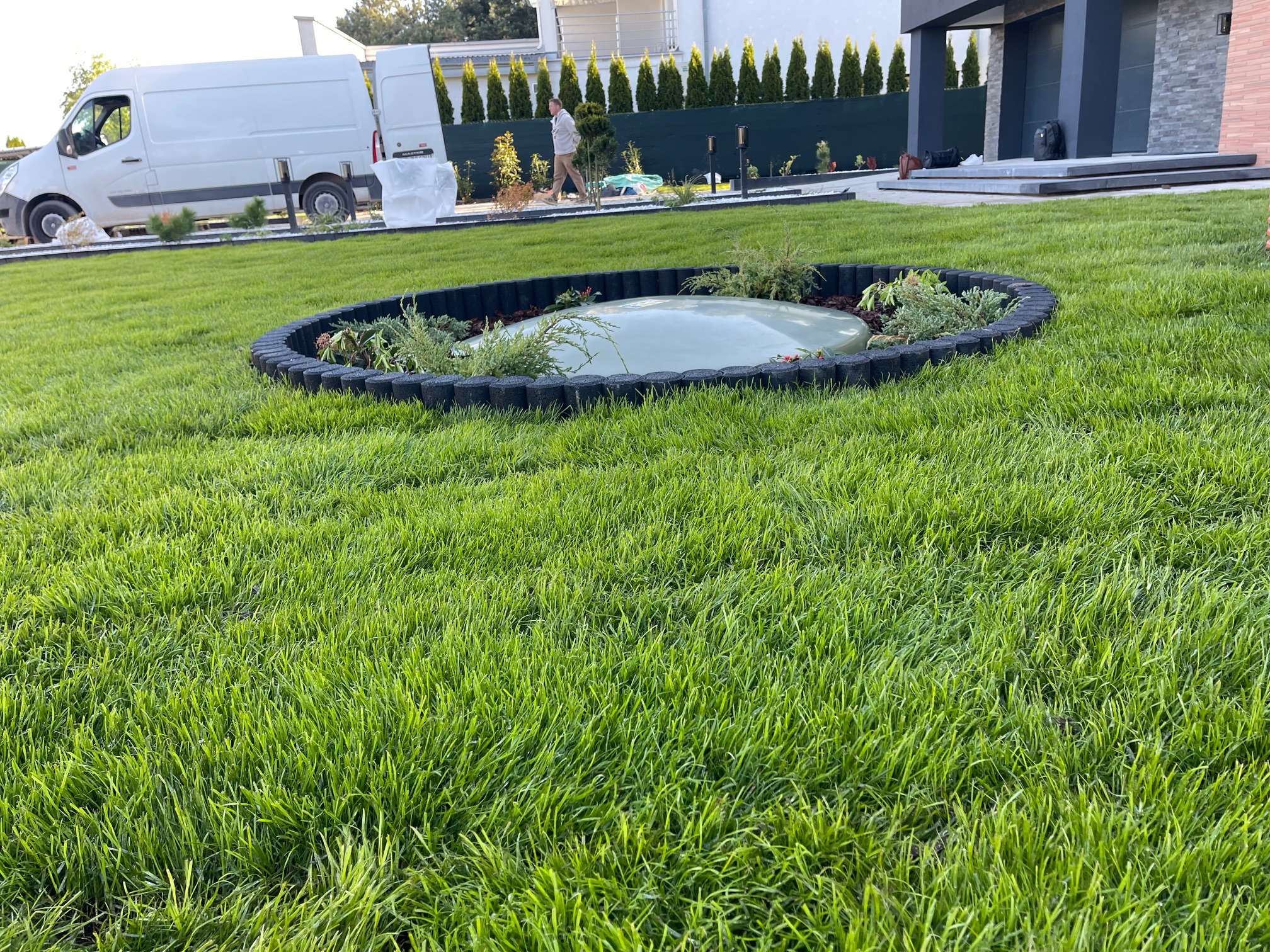 Zadbany trawnik z okrągłym obrzeżem i roślinami, na tle domu i zaparkowanego busa. Widoczny fragment systemu nawadniania.