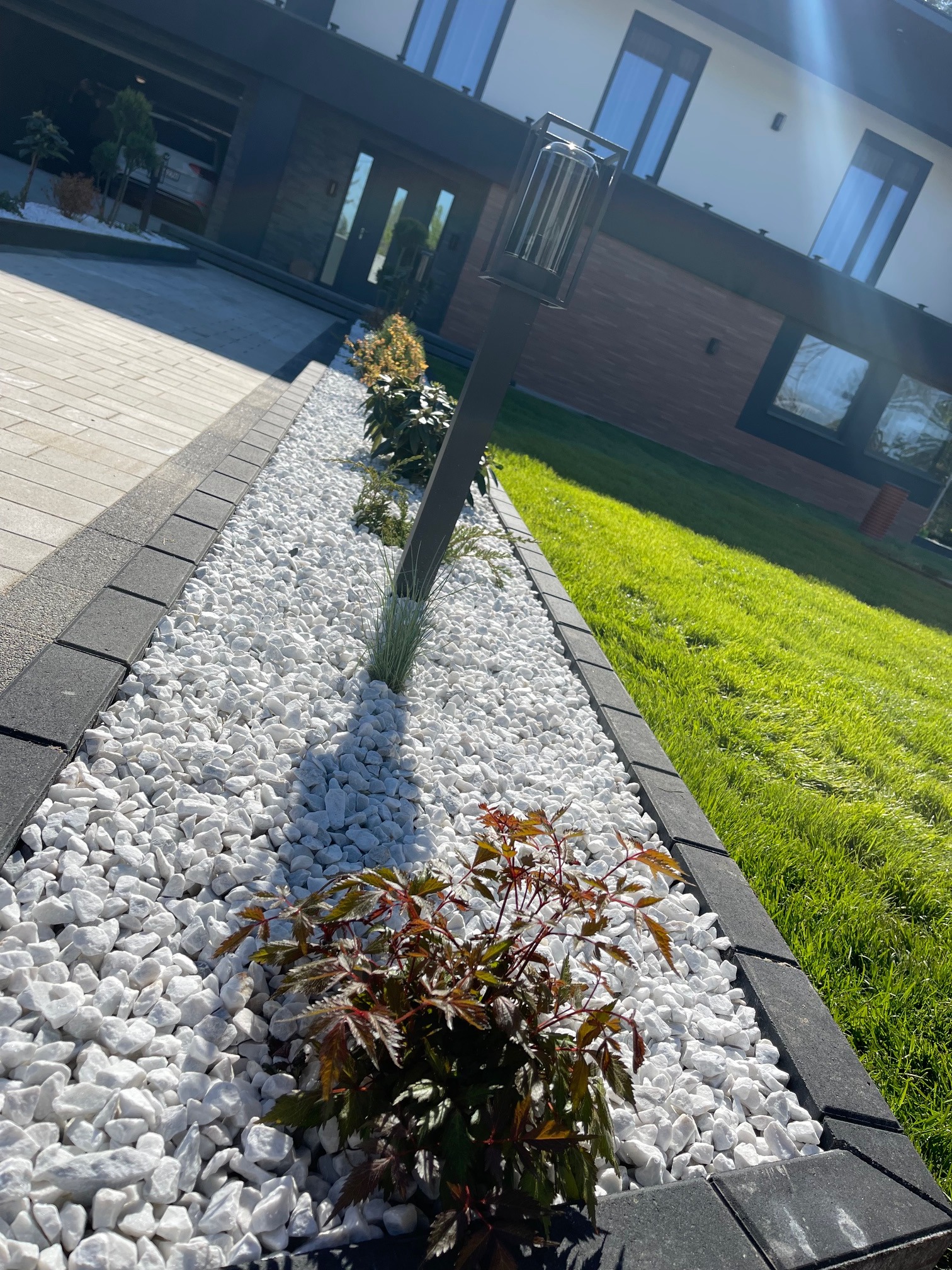 Ogród z białymi kamieniami, niskimi roślinami i lampą wzdłuż ścieżki. Widok na dom z trawnikiem. Elementy małej architektury ogrodowej.