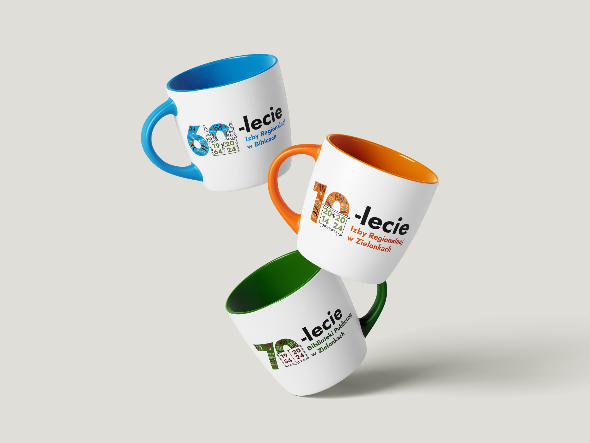 Trzy białe kubki z kolorowymi wnętrzami i uszkami, każdy z innym logo rocznicowym (60-lecie Izby Regionalnej w Bibicach, 10-lecie Izby Regionalnej w Zielonkach, 70-lecie Biblioteki Publicznej...