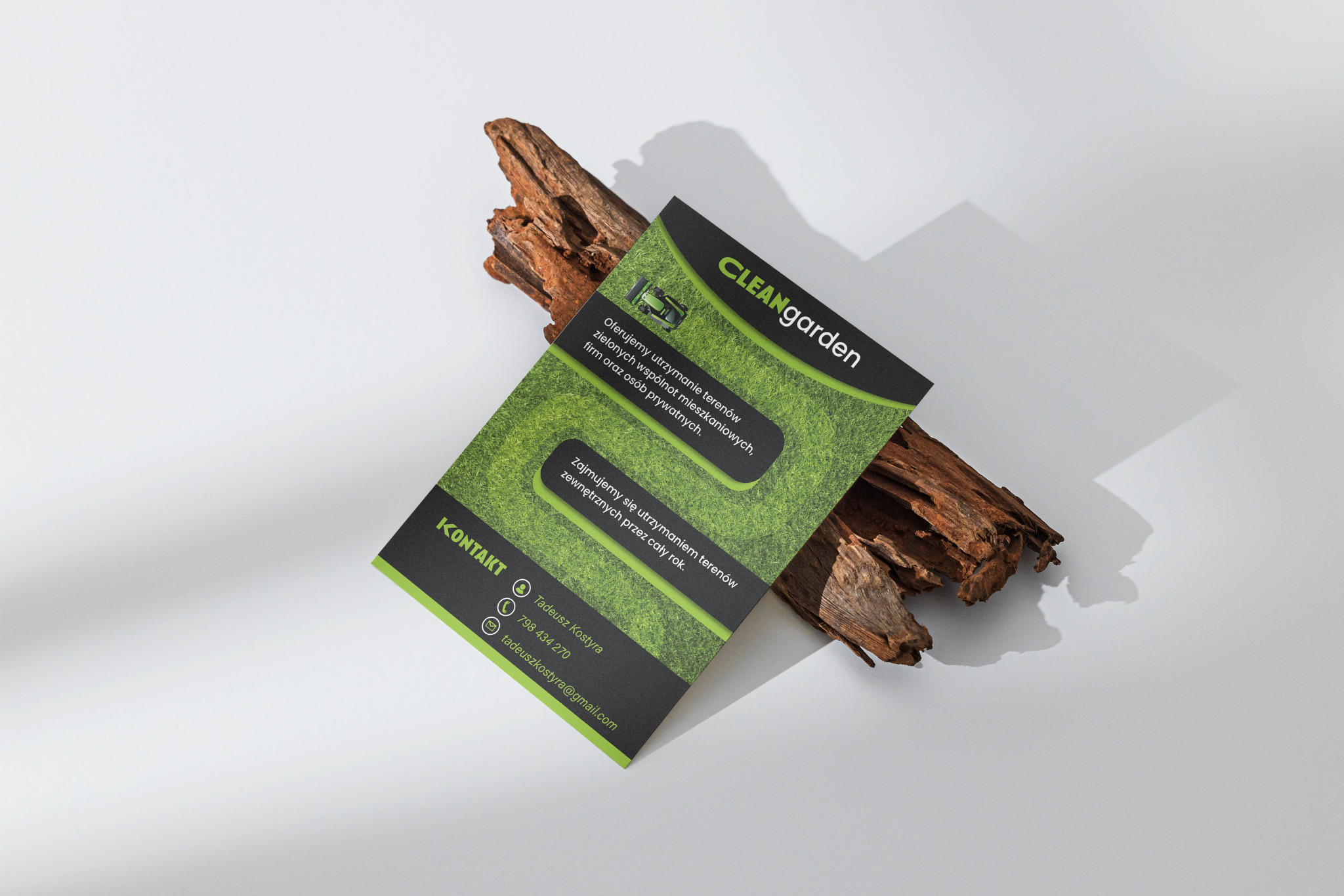 Ulotka reklamowa firmy CleanGarden, specjalizującej się w utrzymaniu terenów zielonych, leżąca na kawałku kory drzewnej, na jasnym tle. Ulotka w kolorach zieleni i czerni, z widocznym logo i danymi...