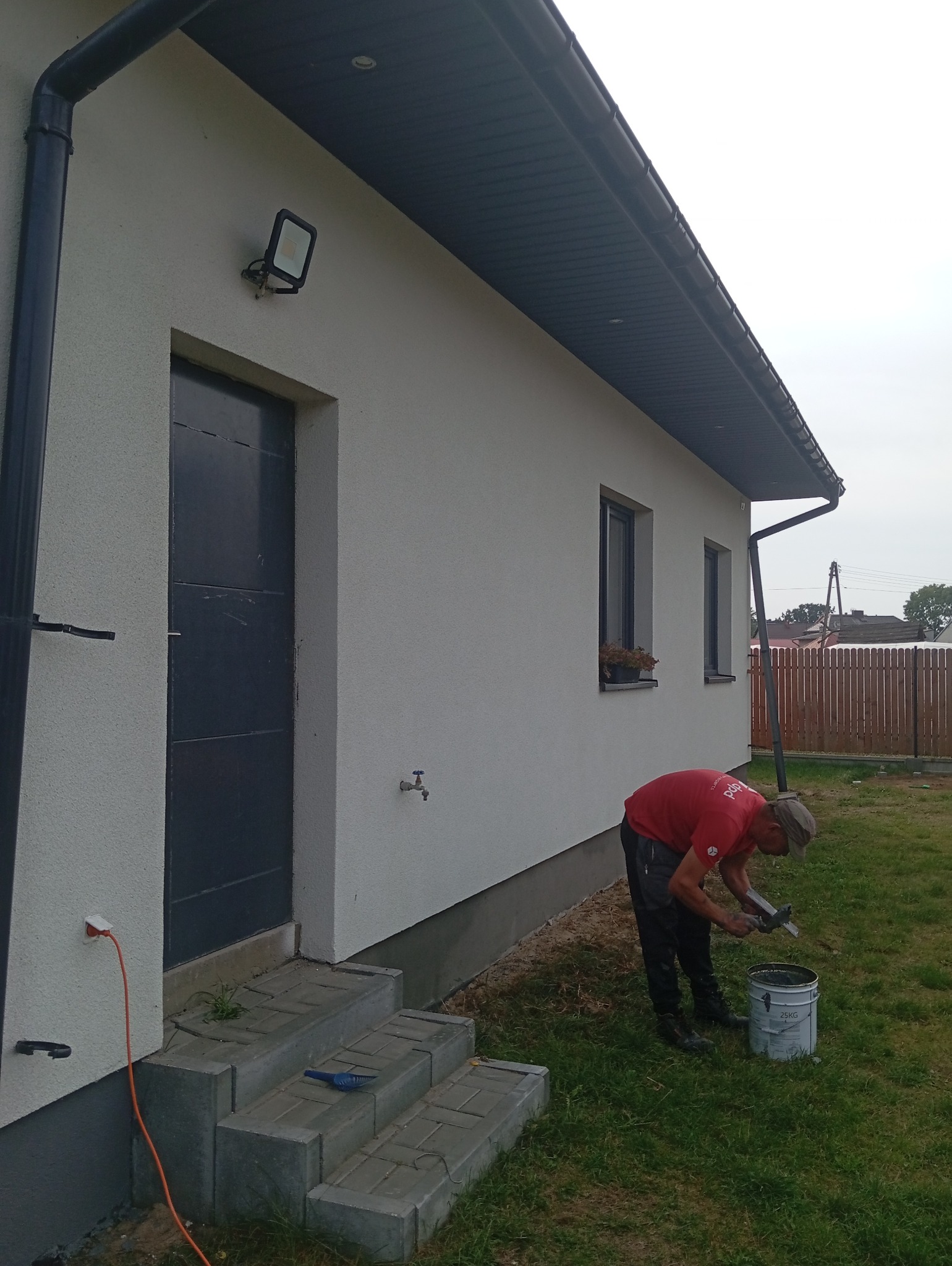 Malowanie elewacji domu jednorodzinnego: mężczyzna w czerwonej koszulce miesza farbę przy użyciu mieszadła, widoczna pomalowana ściana, okna z kwiatami, orynnowanie i lampa elewacyjna.