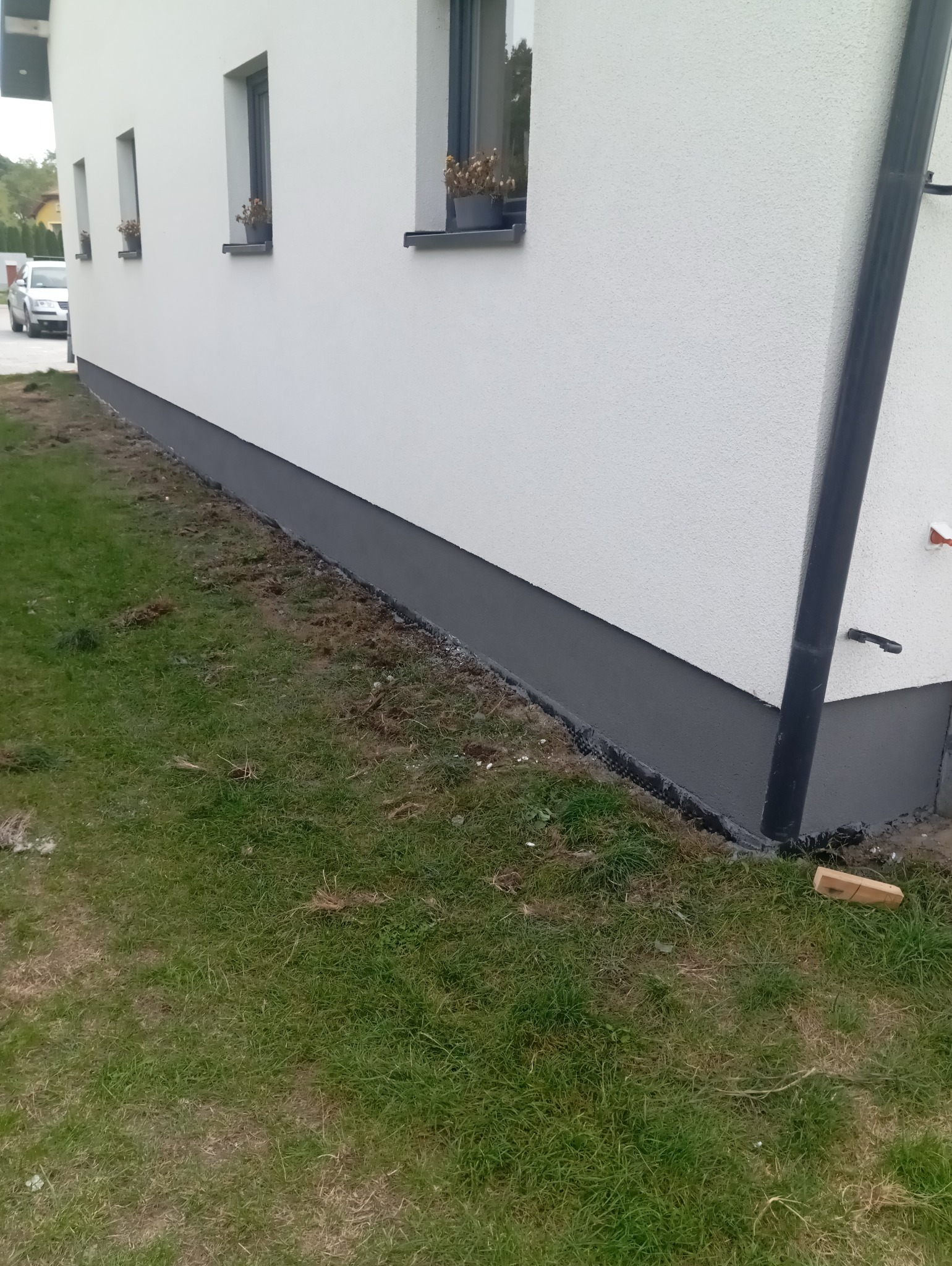 Pomalowana na szaro podmurówka domu jednorodzinnego z białym tynkiem, widoczne okna z kwiatami, rynna spustowa i fragment trawnika.