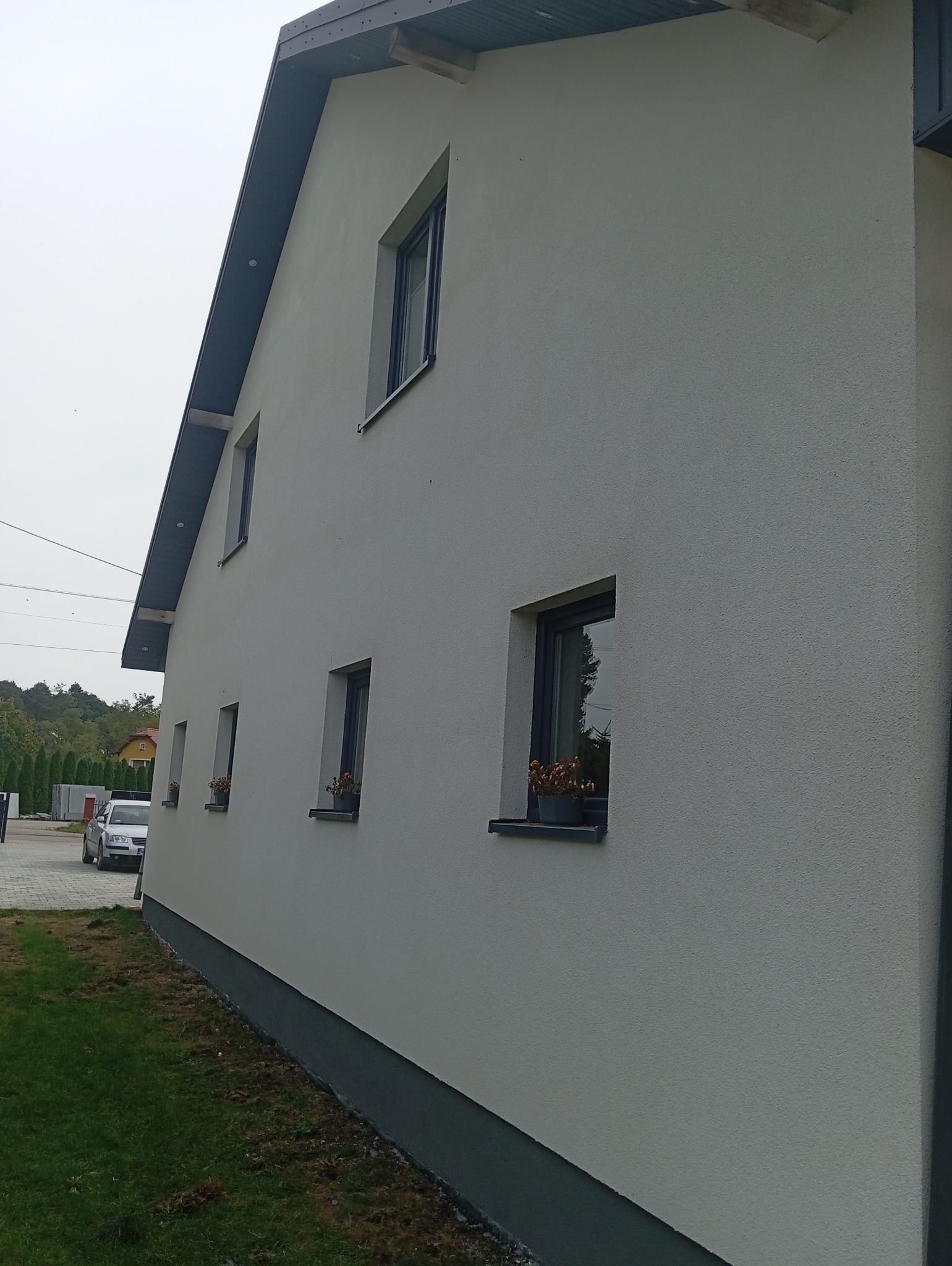 Elewacja domu jednorodzinnego z oknami, na parapetach doniczki z kwiatami, widok z boku, szara podmurówka.