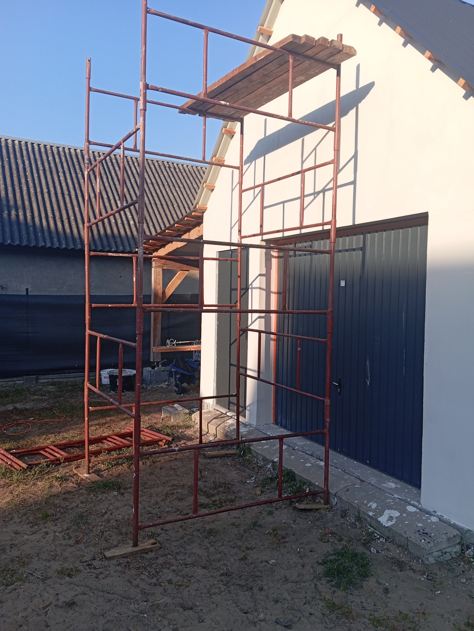 Czerwone rusztowanie ustawione przy świeżo otynkowanej ścianie budynku z szarą bramą garażową i fragmentem dachu widocznym w tle.