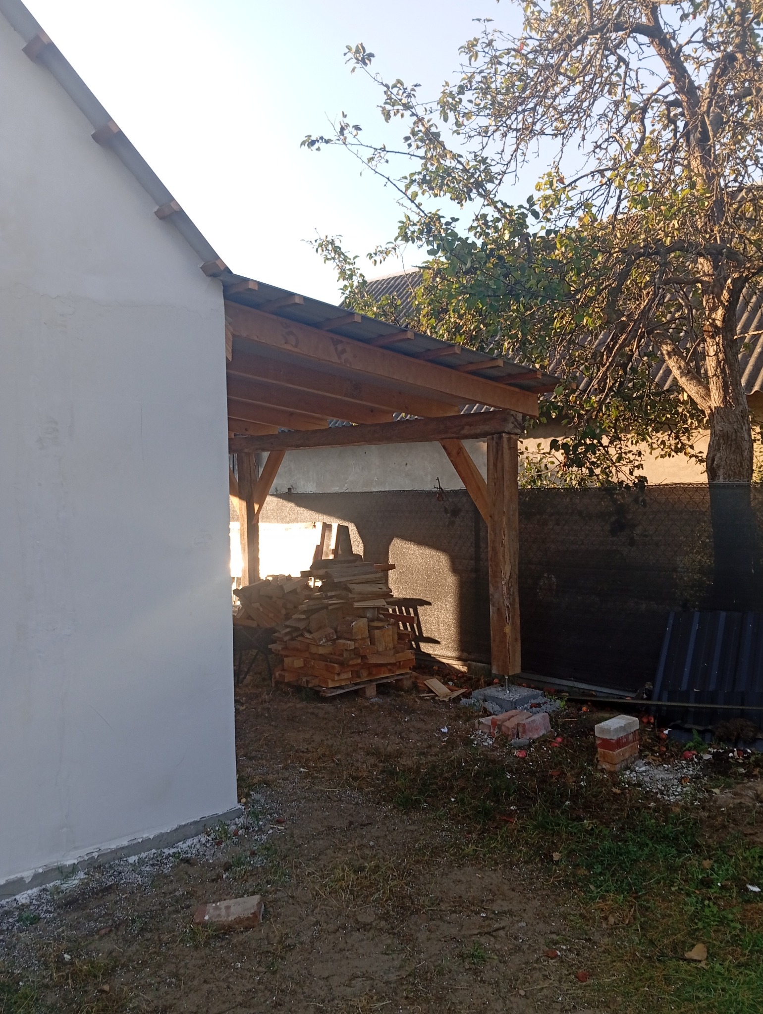 Wznoszenie drewnianej konstrukcji zadaszenia przy budynku z otynkowaną ścianą, widoczne deski, cegły i elementy mocujące, w tle drzewo owocowe.