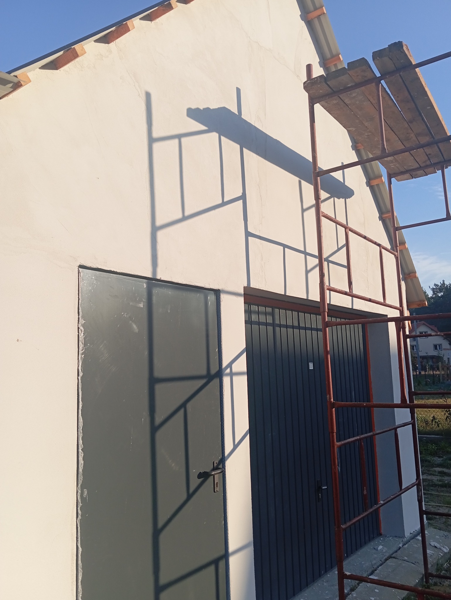 Elewacja budynku po nałożeniu gładzi, widoczne drzwi i brama garażowa, cień rusztowania na ścianie.