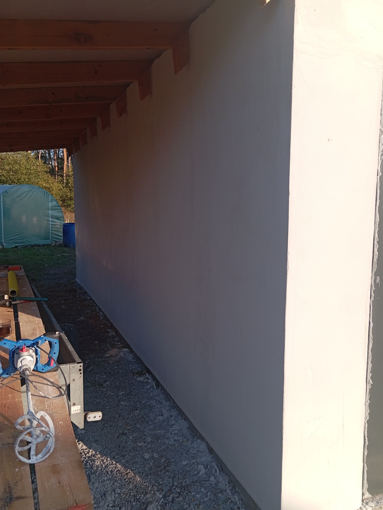 Świeżo otynkowana ściana budynku z widoczną drewnianą konstrukcją stropu, obok stoi mieszadło do zaprawy i narzędzia, w tle widoczny tunel foliowy.