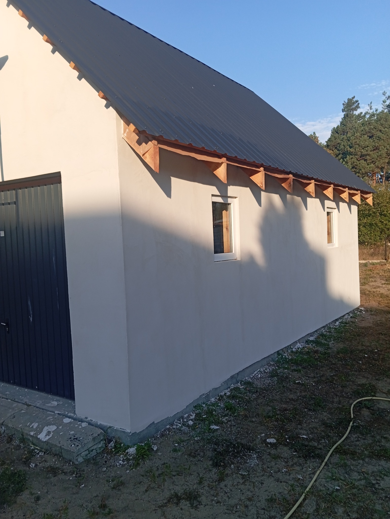 Świeżo otynkowana ściana budynku z szarą blachodachówką, widoczne drewniane elementy pod dachem, okna z białymi ramami i ciemnoszare drzwi garażowe.