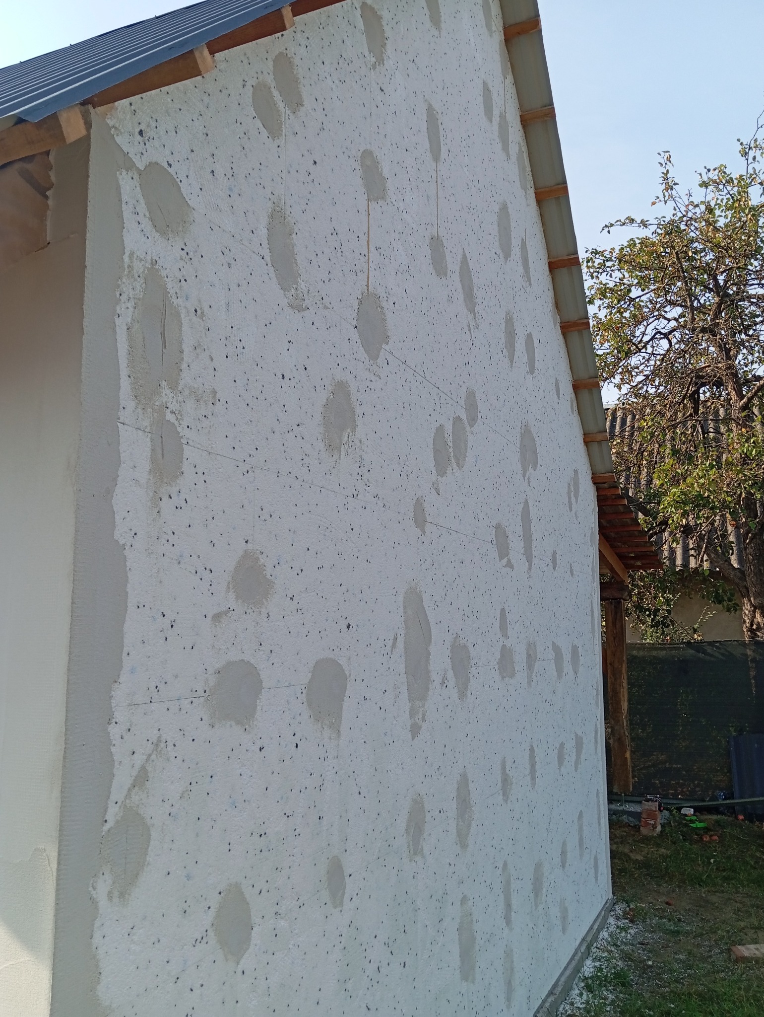 Ocieplona ściana domu w trakcie prac wykończeniowych, widoczne punktowe mocowania styropianu zaprawą klejową, szary dach i drzewo w tle.