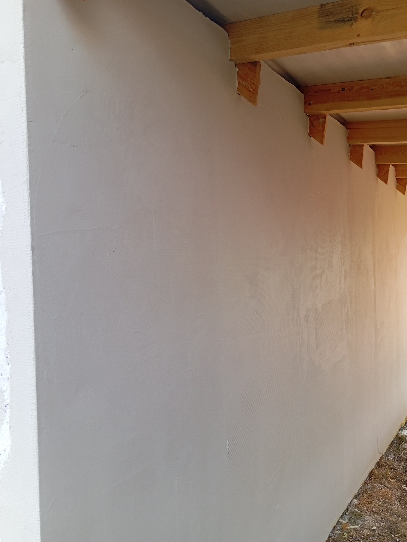 Świeżo położona, jasnoszara gładź na ścianie z widocznymi drewnianymi elementami konstrukcyjnymi pod sufitem, ukazująca etap prac wykończeniowych.