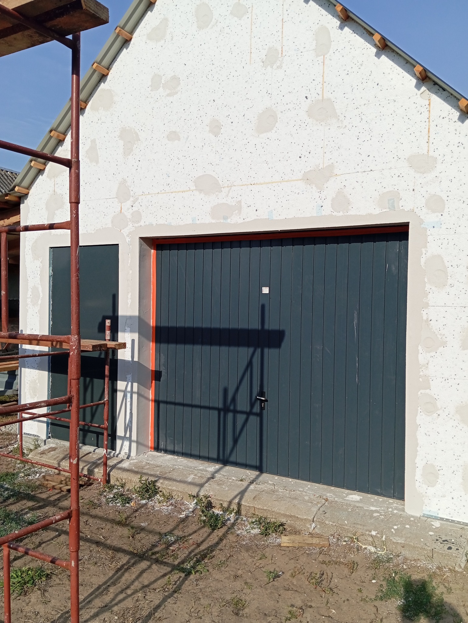 Elewacja budynku w trakcie ocieplania styropianem, widoczne łaty po kleju, szara brama garażowa z pomarańczową ramą, rusztowanie z boku.