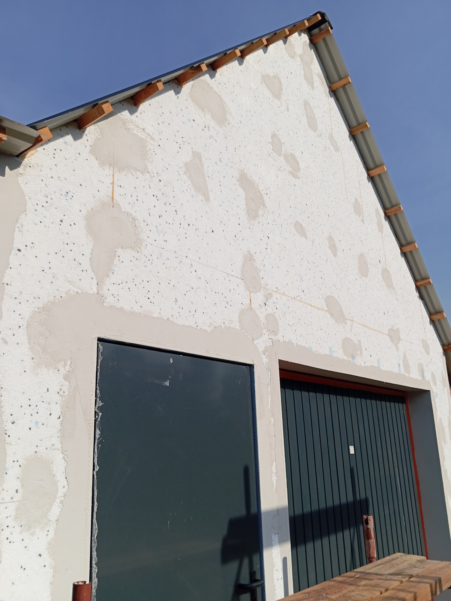 Elewacja budynku w trakcie ocieplania styropianem, z widocznymi łatami po nałożeniu kleju i kołków mocujących; w tle błękitne niebo.