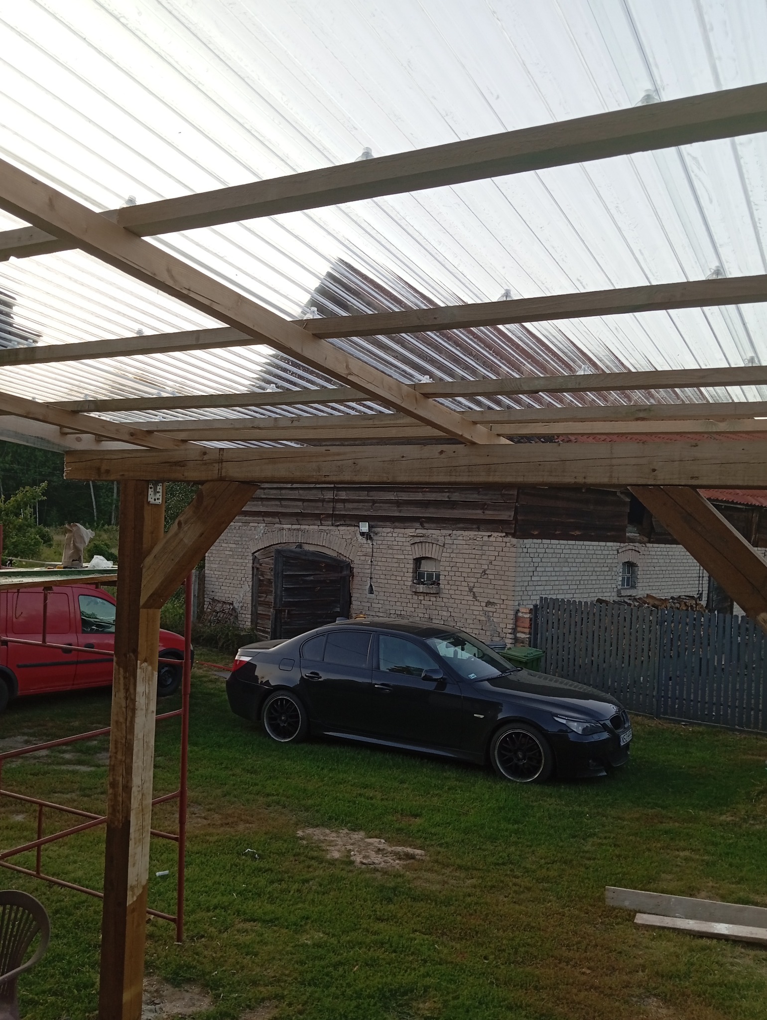 Konstrukcja drewnianej wiaty z przezroczystym zadaszeniem poliwęglanowym, widoczny fragment czerwonego samochodu i czarny samochód osobowy na trawniku, w tle budynek gospodarczy z cegły.