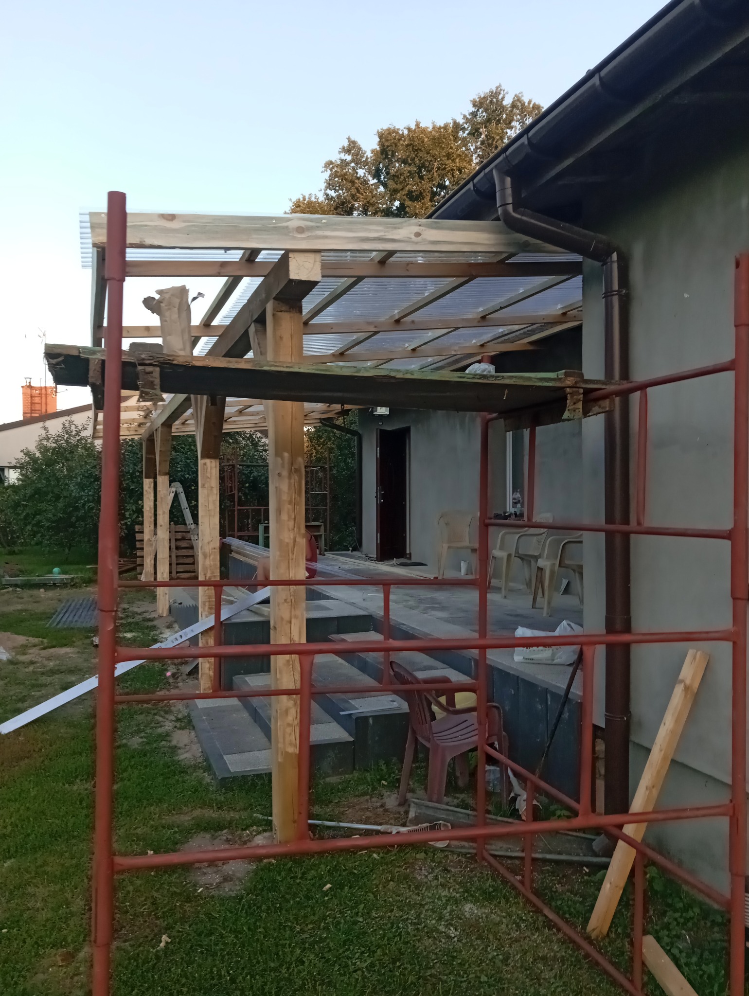 Budowa zadaszenia tarasu z użyciem drewnianej konstrukcji i przezroczystego pokrycia, widoczne rusztowanie i elementy wykończeniowe.