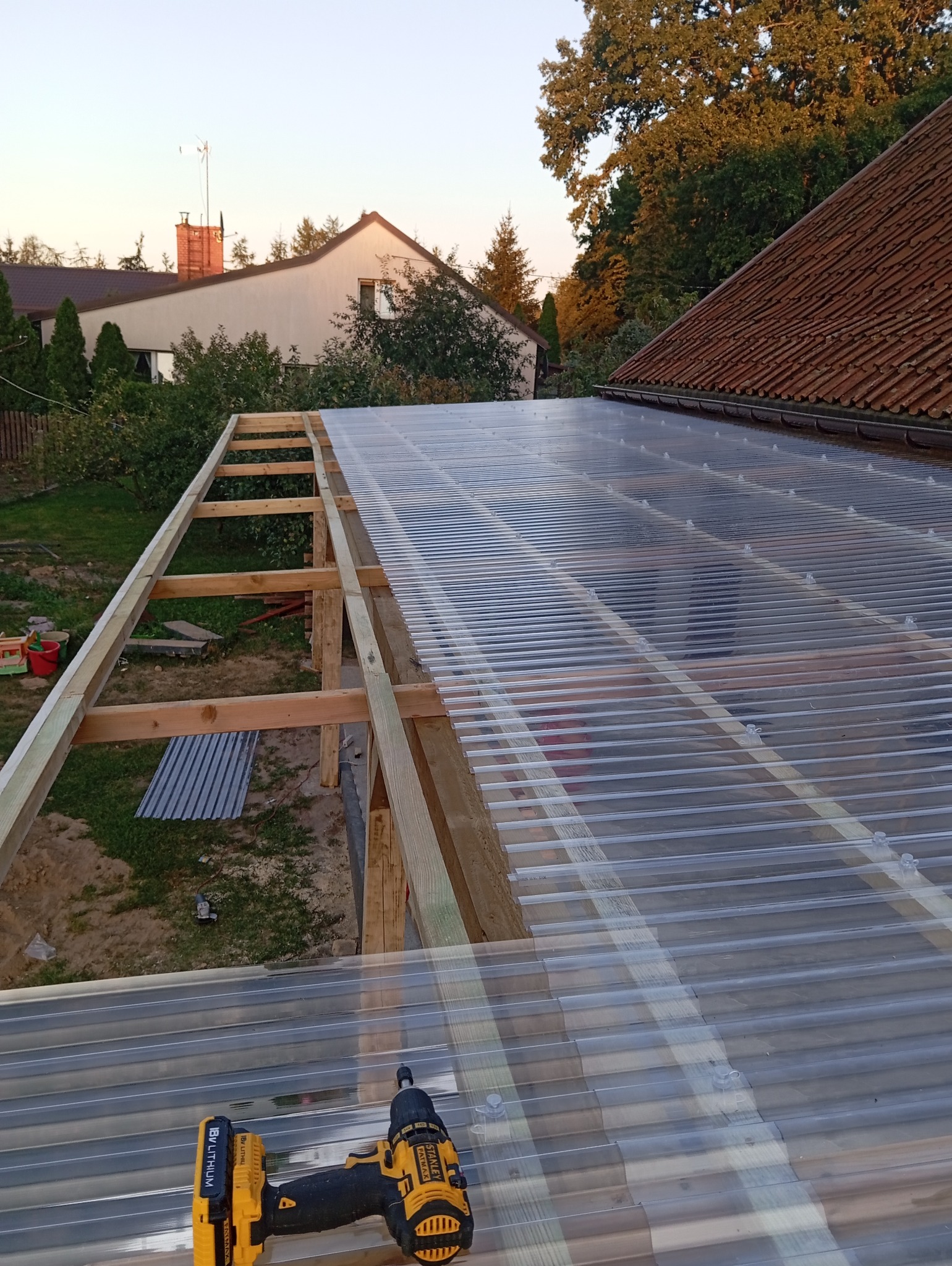 Montaż przezroczystego zadaszenia z poliwęglanu falistego na drewnianej konstrukcji, w tle budynek mieszkalny, widoczna wkrętarka akumulatorowa.