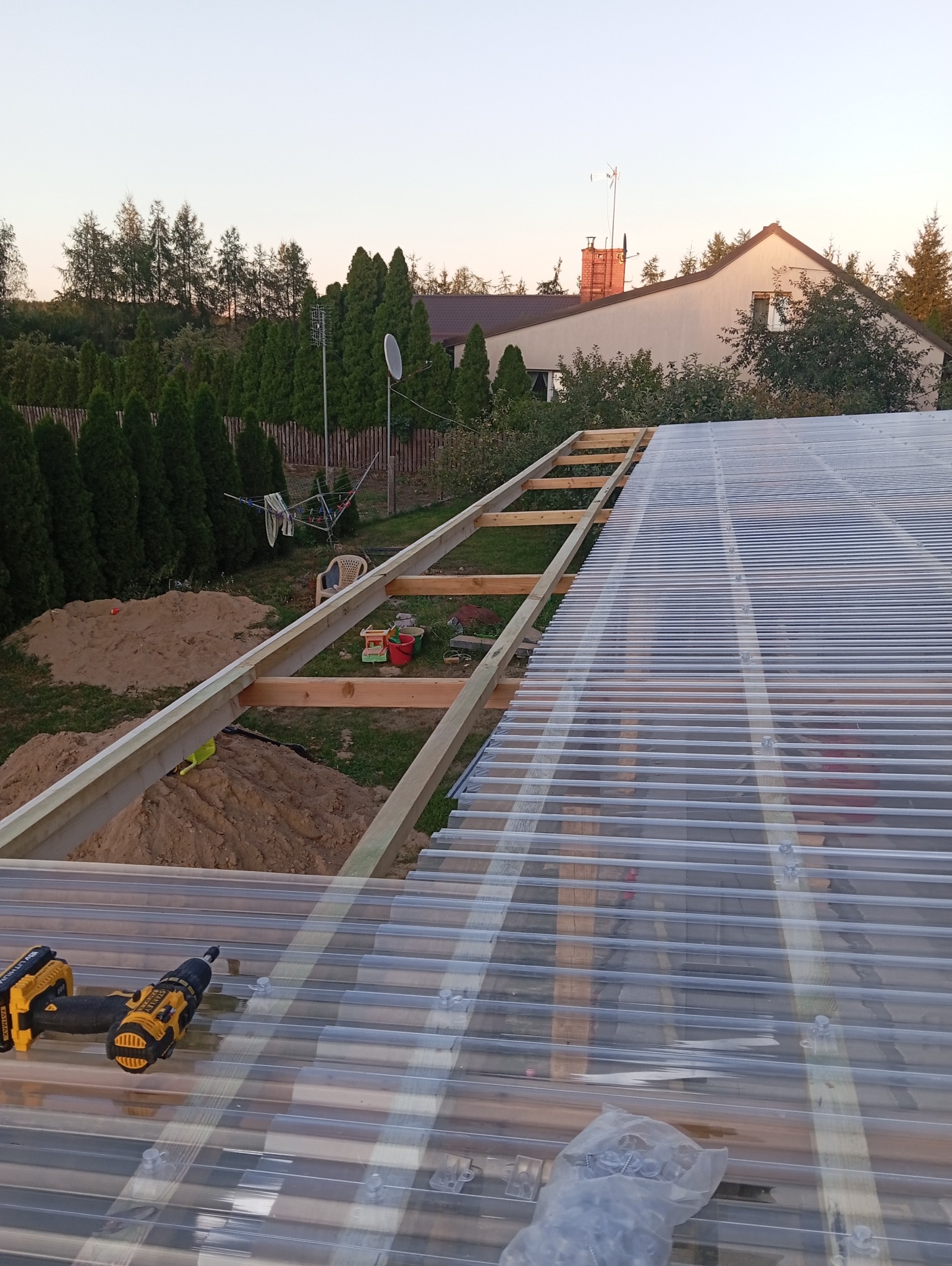 Montaż przezroczystego zadaszenia z poliwęglanu falistego na drewnianej konstrukcji, widoczne wkrętarki akumulatorowe i worek ze śrubami, w tle ogród z domem.