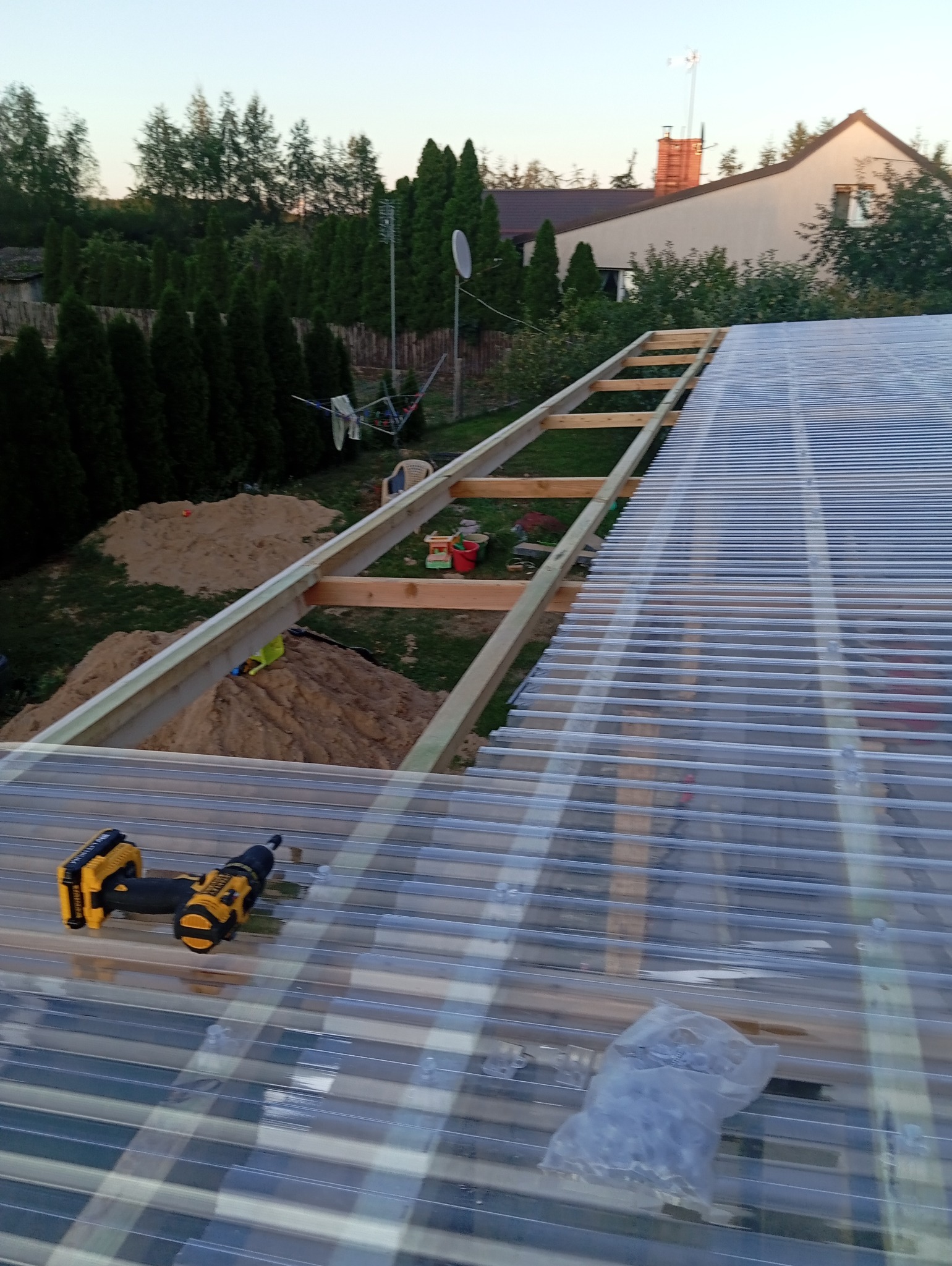 Montaż przezroczystego zadaszenia z poliwęglanu falistego na drewnianej konstrukcji, widoczne wkrętarki akumulatorowe, worek ze śrubami, w tle ogród z tujami i dom