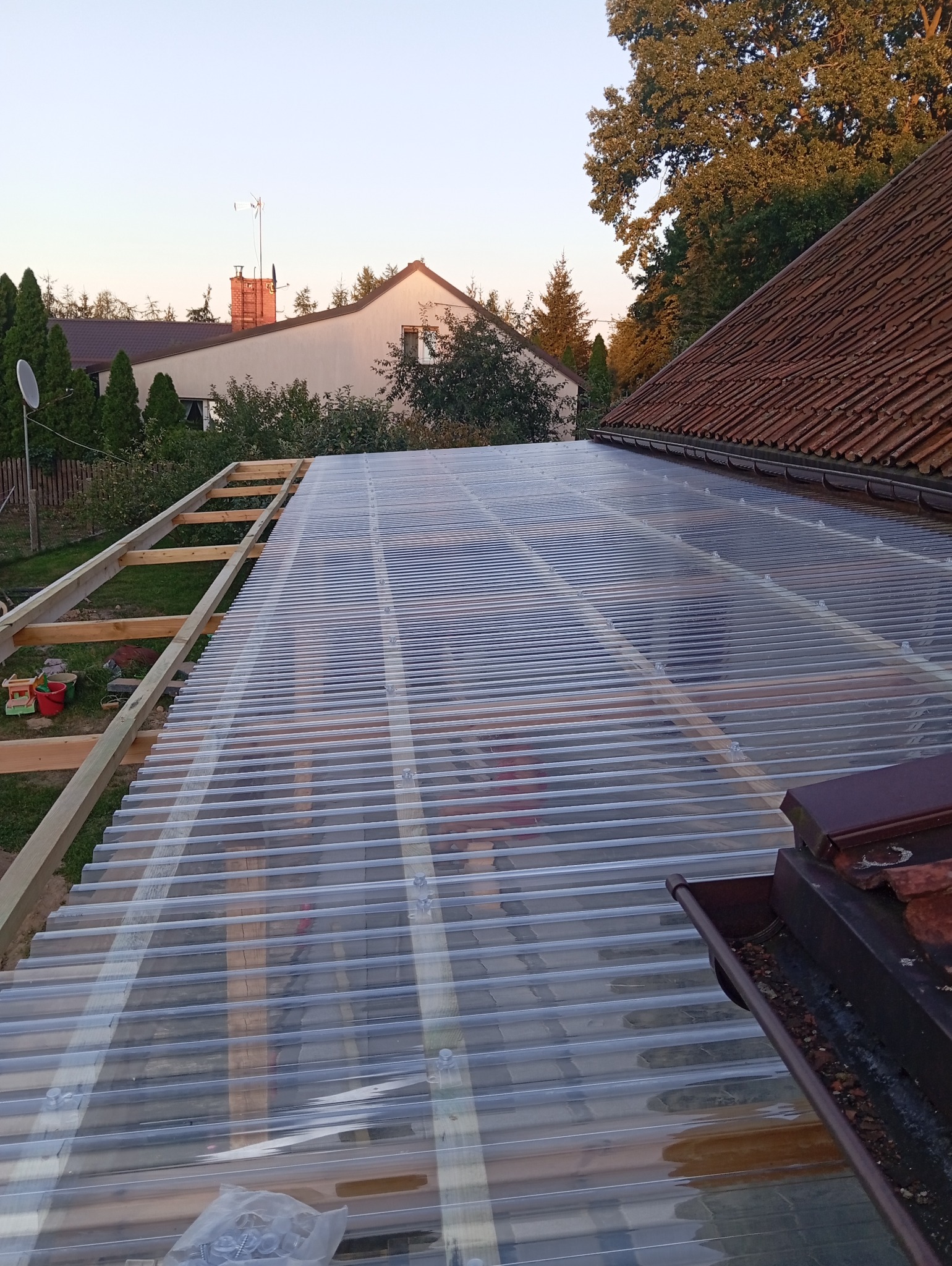 Konstrukcja z drewna i transparentnego poliwęglanu falistego tworząca zadaszenie tarasu w trakcie montażu, widok z góry na tle domów i zieleni.