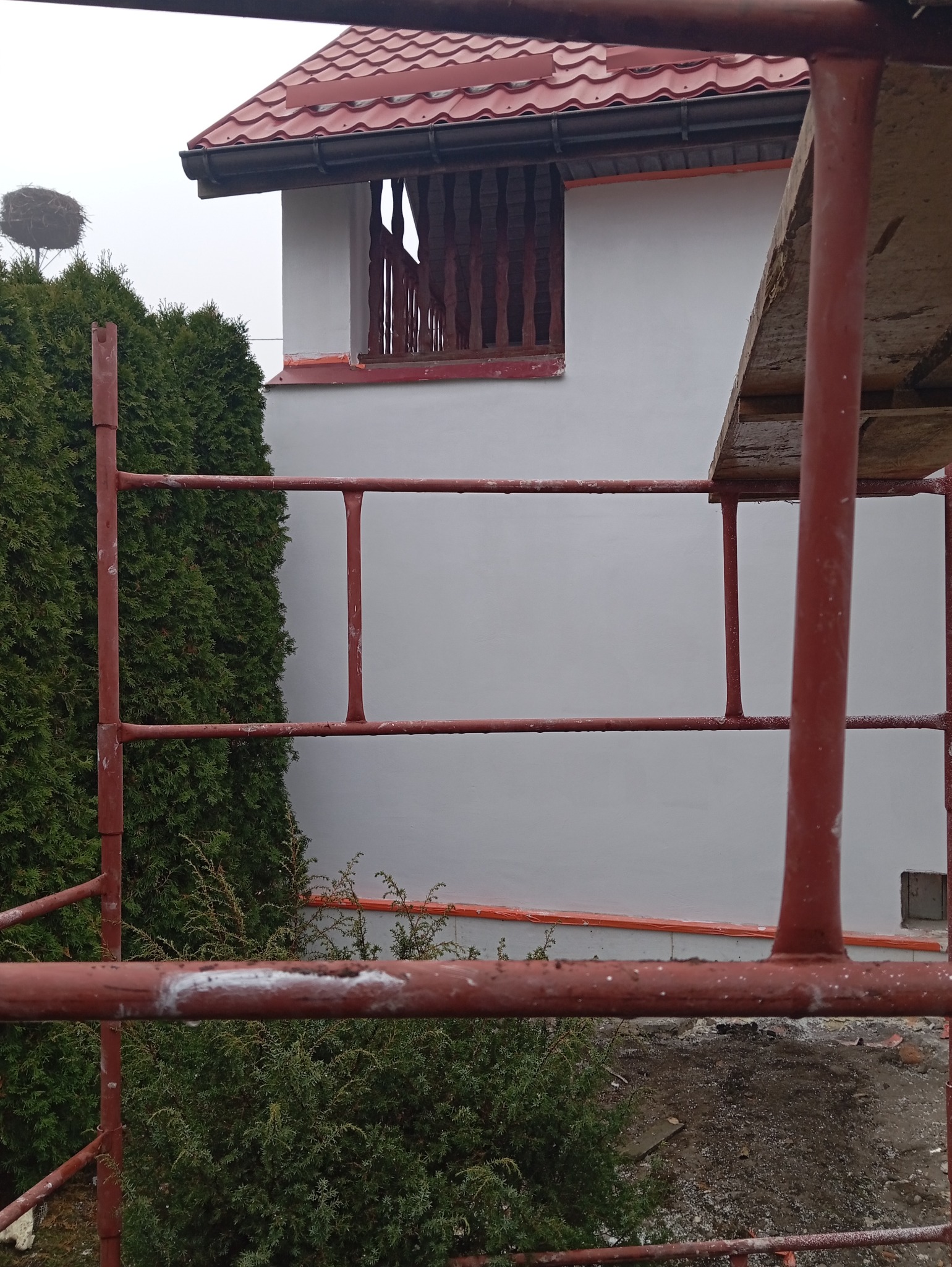 Dom w trakcie malowania elewacji, widoczne rusztowanie, świeżo pomalowana ściana na szaro z czerwonymi akcentami, dach z czerwonej dachówki i gniazdo bocianie na drzewie w tle.