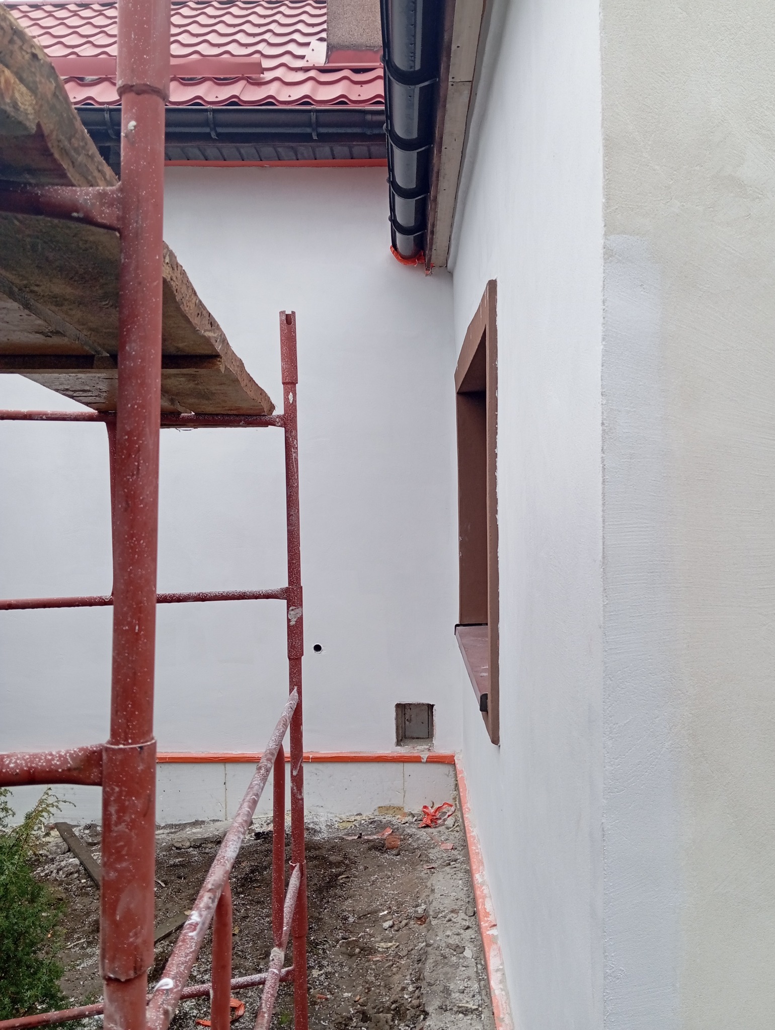 Świeżo otynkowana elewacja budynku z widocznym fragmentem rusztowania, zabezpieczeniem okna i otworu w ścianie, podkreślającym trwający remont domu.