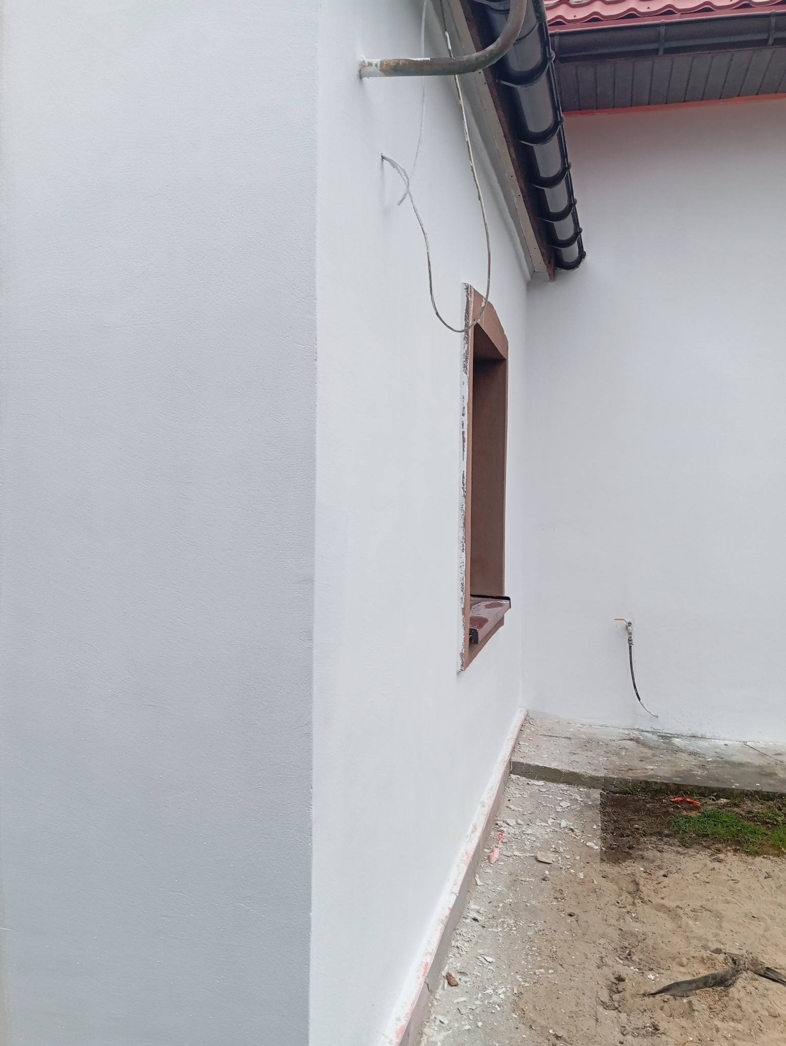 Boczny widok na fragment elewacji domu po remoncie, z oknem bez szyby, świeżo pomalowaną białą ścianą, widoczną rurą od instalacji i fragmentem dachu z rynną.