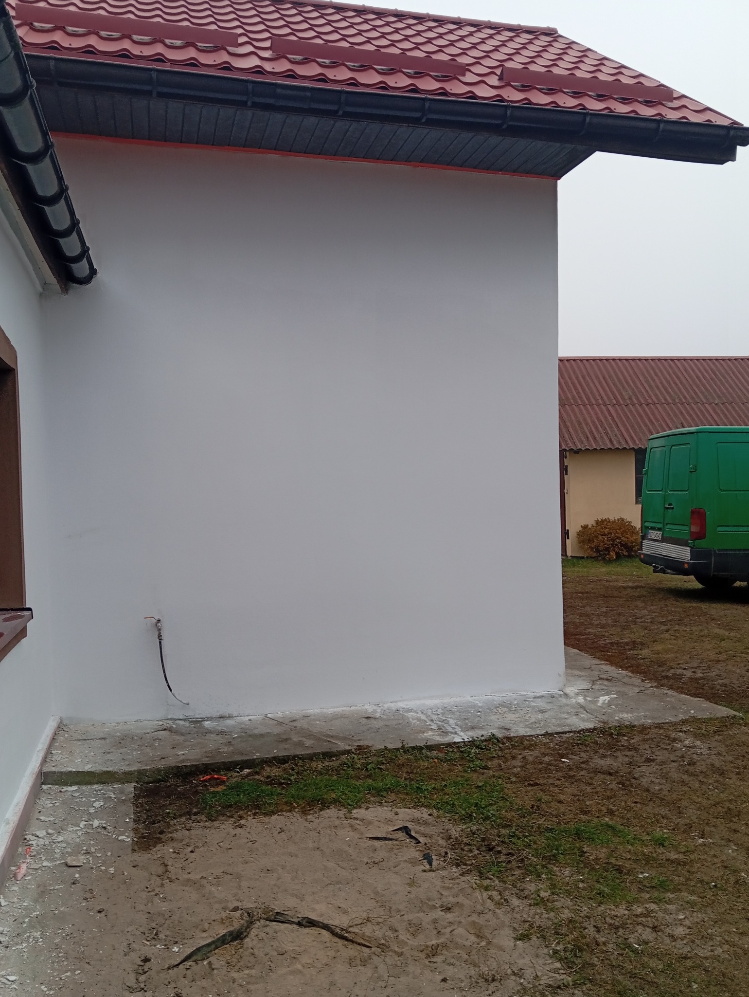 Świeżo pomalowana ściana zewnętrzna budynku z czerwoną dachówką, widoczny fragment okna, rura gazowa i zielony samochód dostawczy w tle.