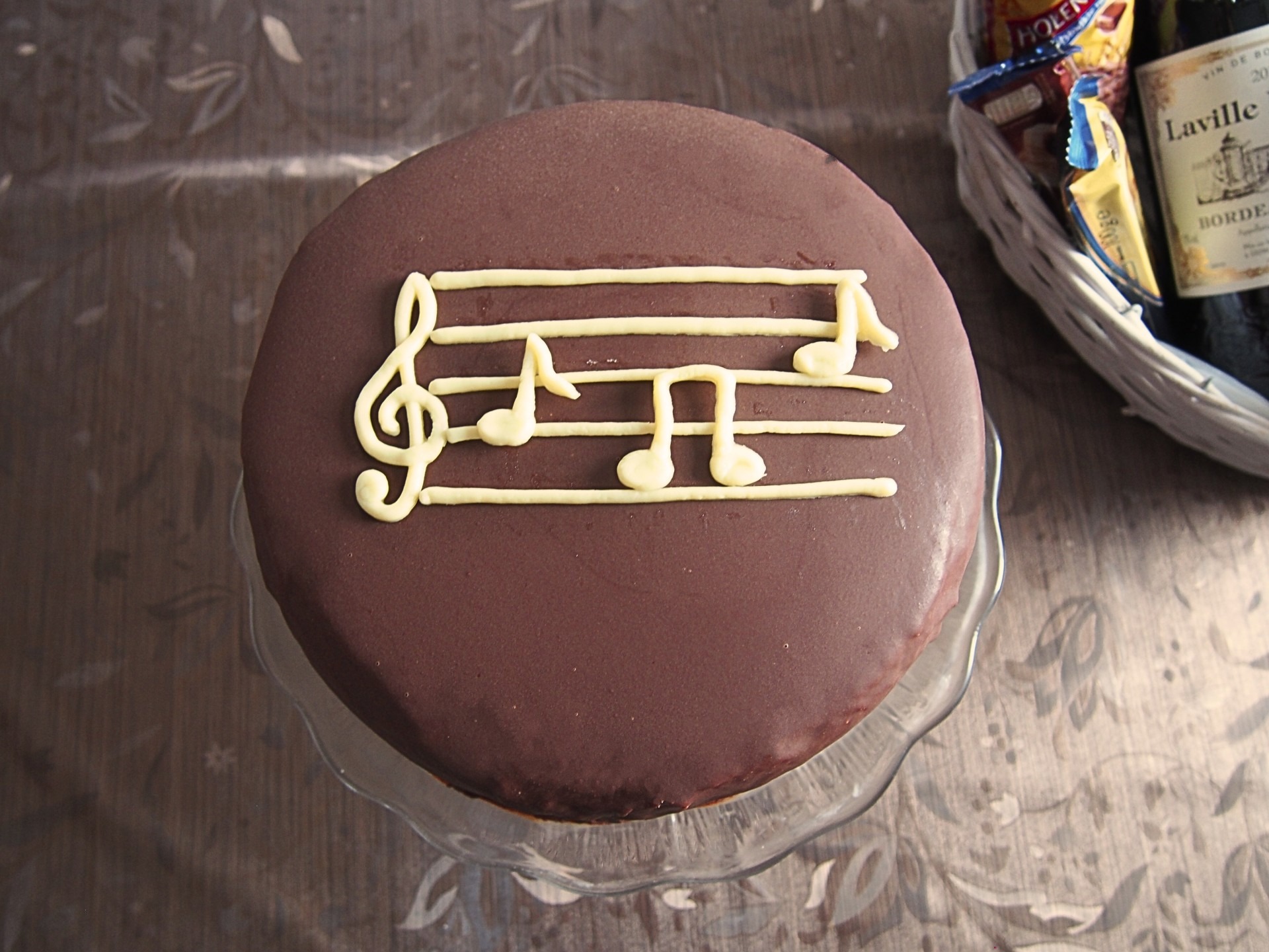 Tort czekoladowy na szklanej paterze, udekorowany nutami z białej czekolady, obok koszyk z przekąskami i butelką wina.