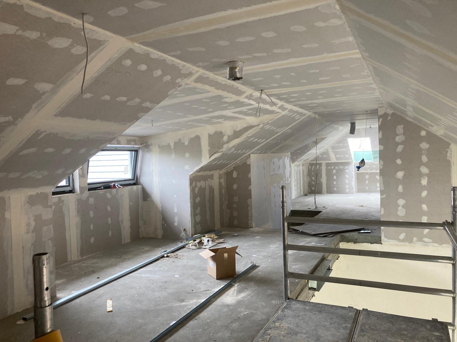 Poddasze w trakcie wykańczania: widoczne płyty gipsowo-kartonowe na ścianach i suficie, zaszpachlowane łączenia, metalowe profile na podłodze, kartonowe pudełko i rusztowanie w pomieszczeniu...