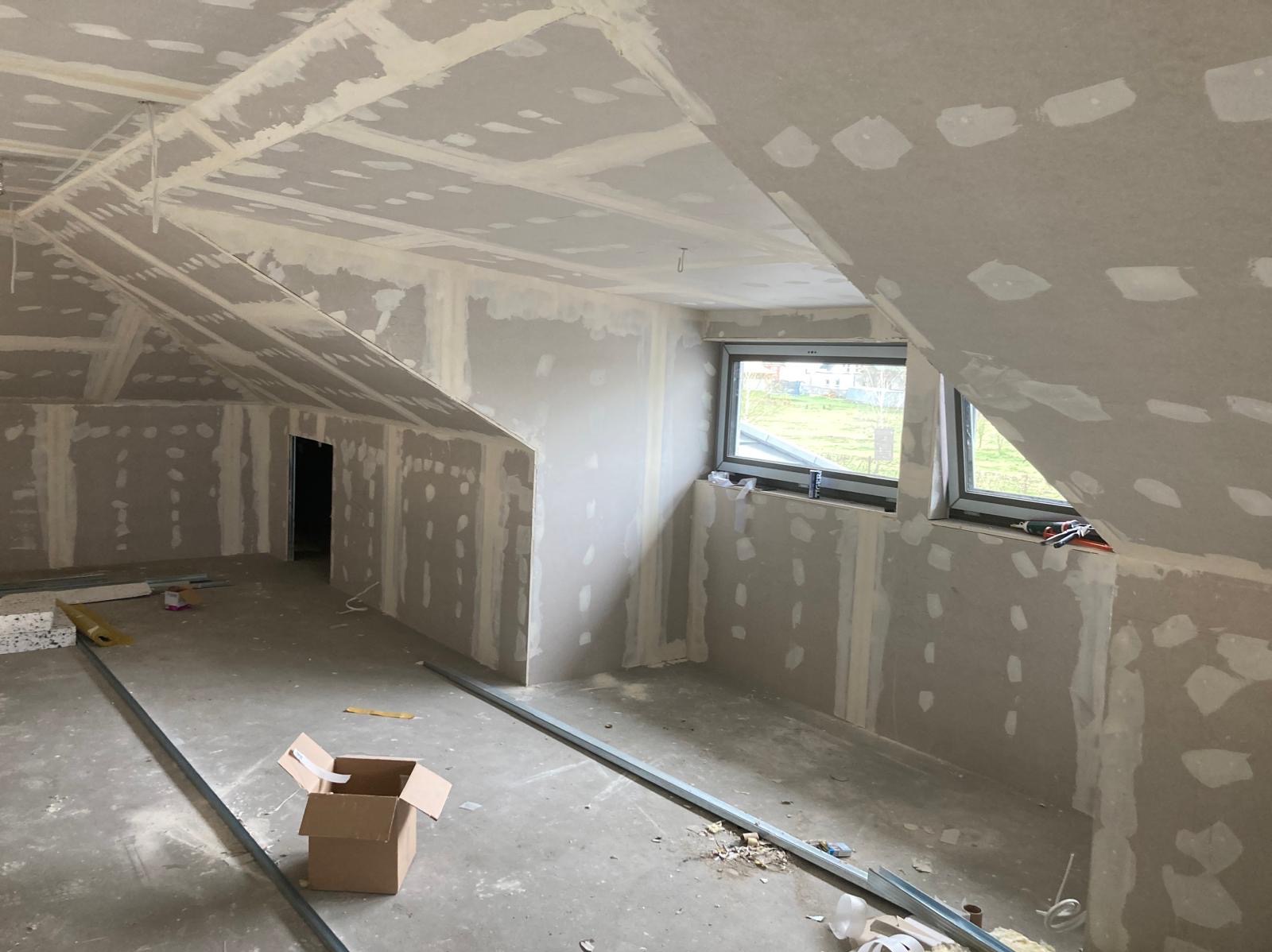 Poddasze w trakcie wykańczania płytami gipsowo-kartonowymi, widoczne szpachlowane łączenia, metalowe profile na podłodze, kartonowe pudło i okna dachowe z widokiem na zieleń.