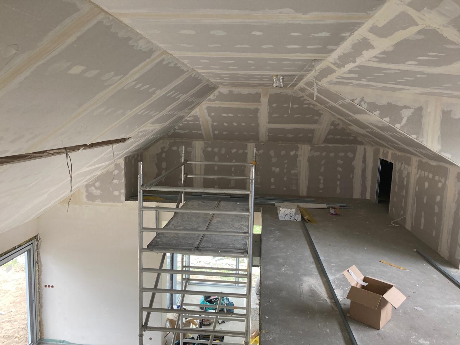 Poddasze w trakcie wykańczania, widoczne szpachlowane łączenia płyt na skośnym suficie i ścianach, metalowe profile, rusztowanie i karton na podłodze.