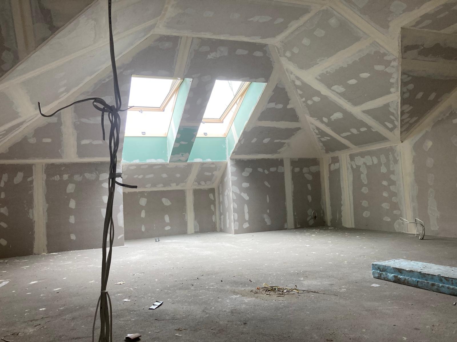 Poddasze w trakcie wykańczania: ściany i skośny sufit obłożone płytami, widoczne łączenia szpachlowane, dwa okna dachowe w zielonej wnęce, przewody elektryczne zwisające z sufitu, surowa podłoga.