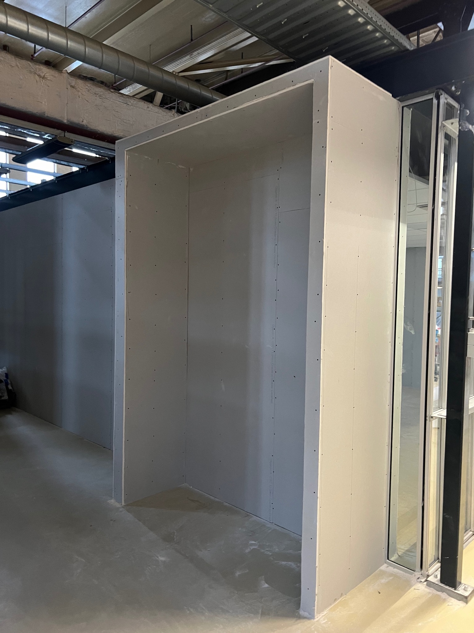 Niedokończona konstrukcja z płyt gipsowo-kartonowych tworząca wnękę w pomieszczeniu komercyjnym, widoczne ślady szpachlowania na łączeniach, surowy beton na podłodze, instalacje pod sufitem.