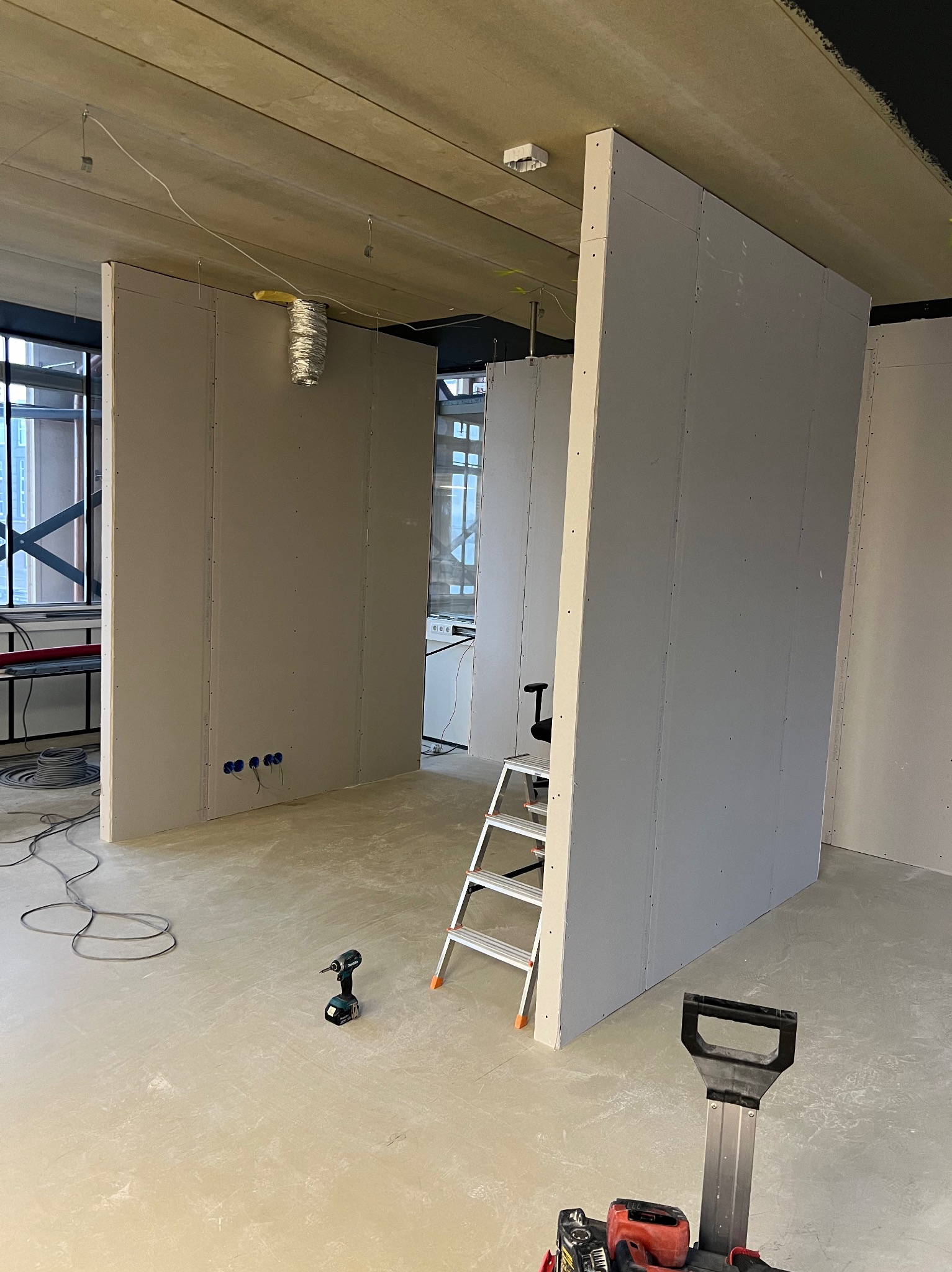 Wnętrze w trakcie prac wykończeniowych: pionowe ścianki działowe z płyt gipsowo-kartonowych, widoczne narzędzia (wiertarka, szlifierka), drabina aluminiowa, surowy betonowy sufit z instalacją...