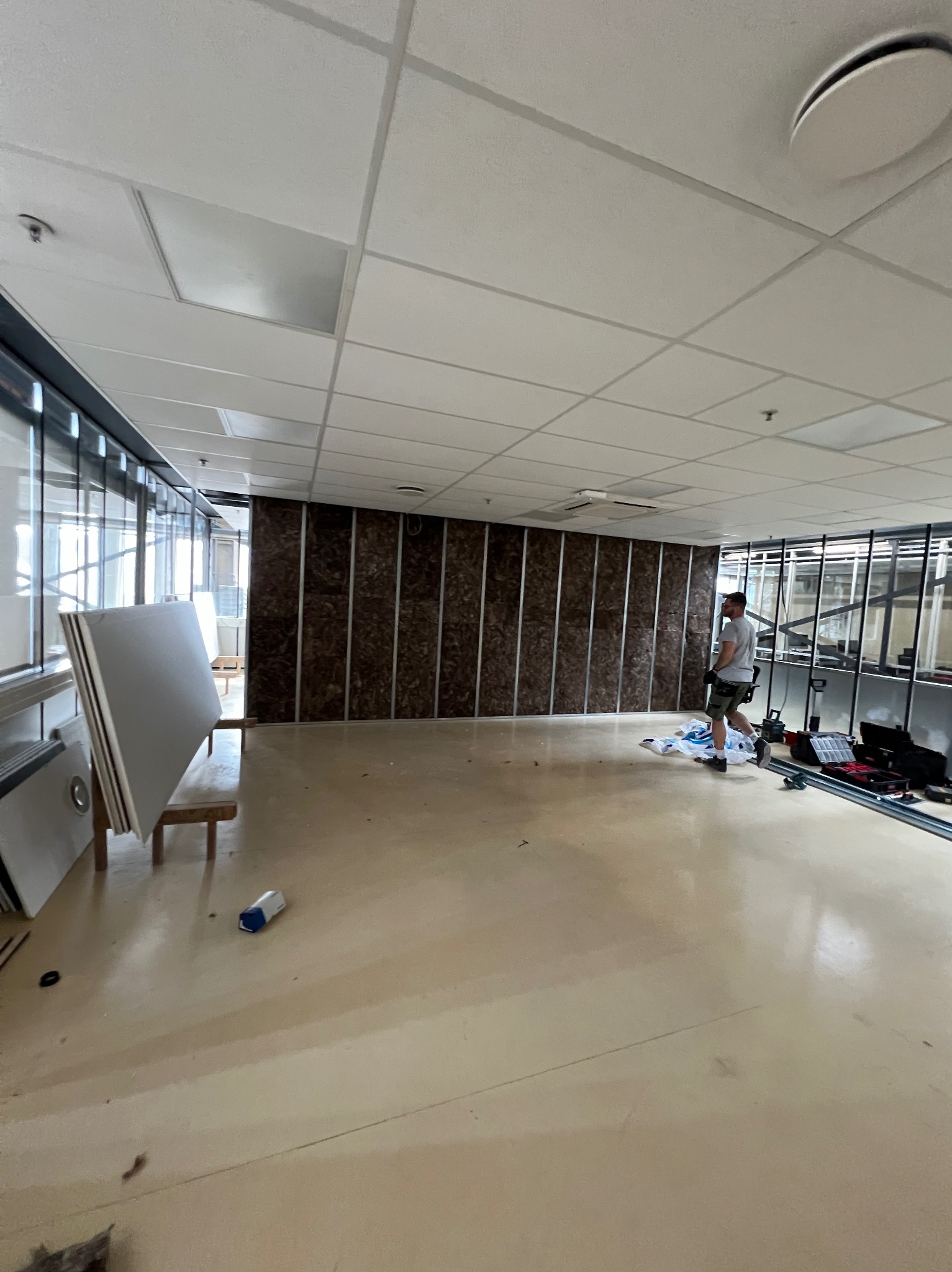 Montaż izolacji na ścianie z metalowym stelażem, widoczne płyty karton gips na drewnianym stojaku i narzędzia pracy rozłożone na podłodze w jasnym, przestronnym pomieszczeniu biurowym.