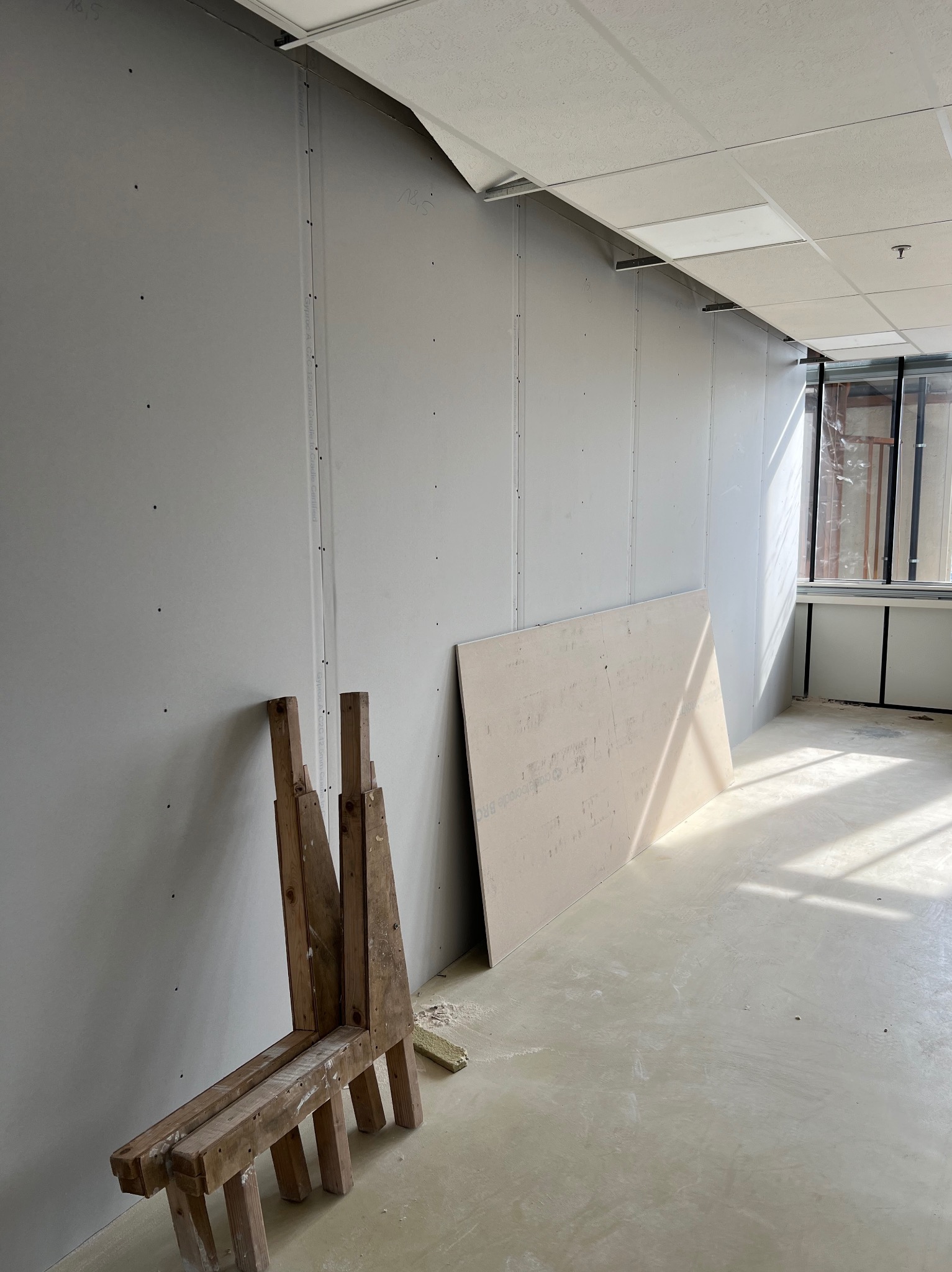 Wnętrze w trakcie montażu ścian działowych z płyt gipsowo-kartonowych, widoczne łączenia płyt, drewniany kozioł i nieuprzątnięty bałagan na podłodze.