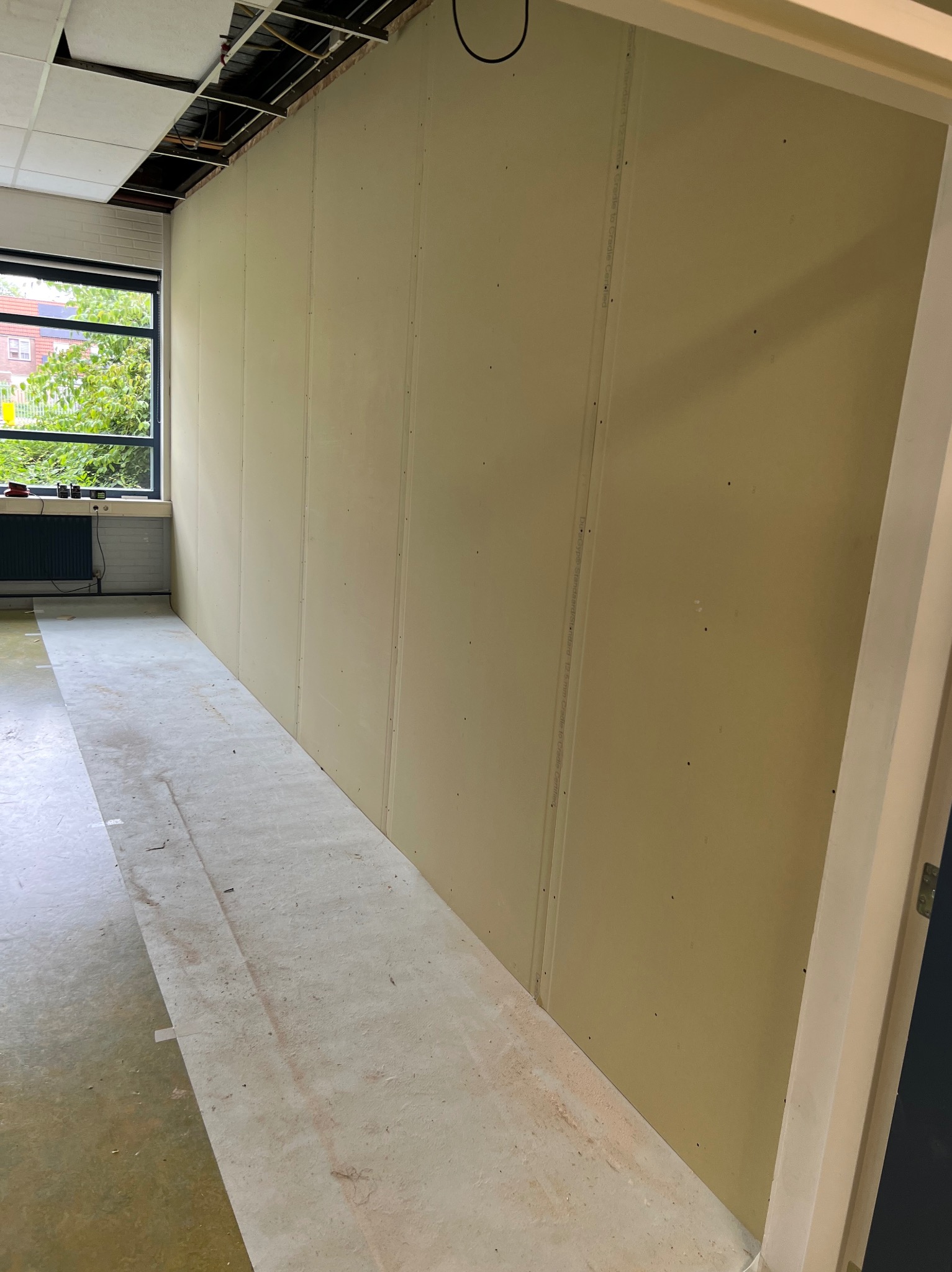 Wnętrze w trakcie remontu: zamontowane płyty gipsowo-kartonowe na ścianie, zabezpieczona podłoga, widoczne okno i fragment sufitu podwieszanego w trakcie montażu.