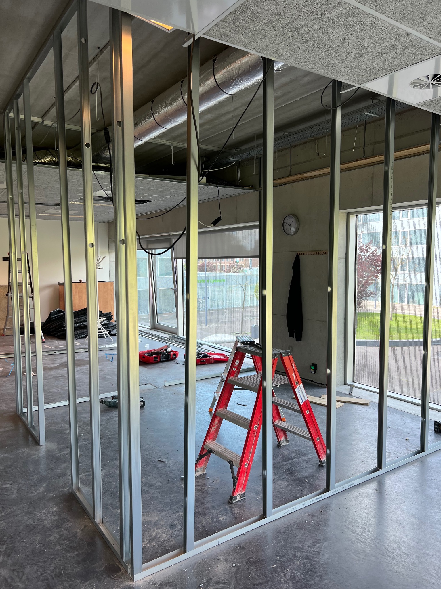 Metalowa konstrukcja ścian działowych w biurze, widoczna czerwona drabina, narzędzia i okna z widokiem na budynek.
