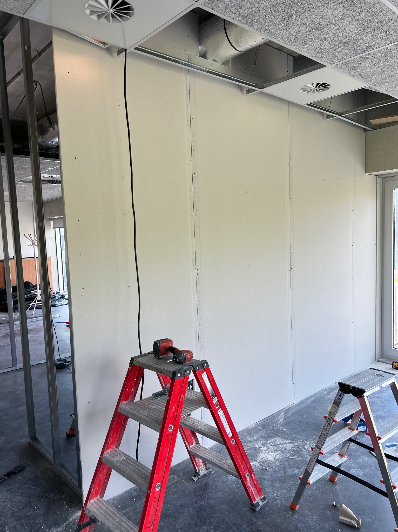 Wnętrze w trakcie wykańczania: zamontowane płyty na ścianie, widoczne profile metalowe konstrukcji, czerwona drabina z wkrętarką na górnym stopniu, druga drabina obok, sufit z widocznymi elementami...