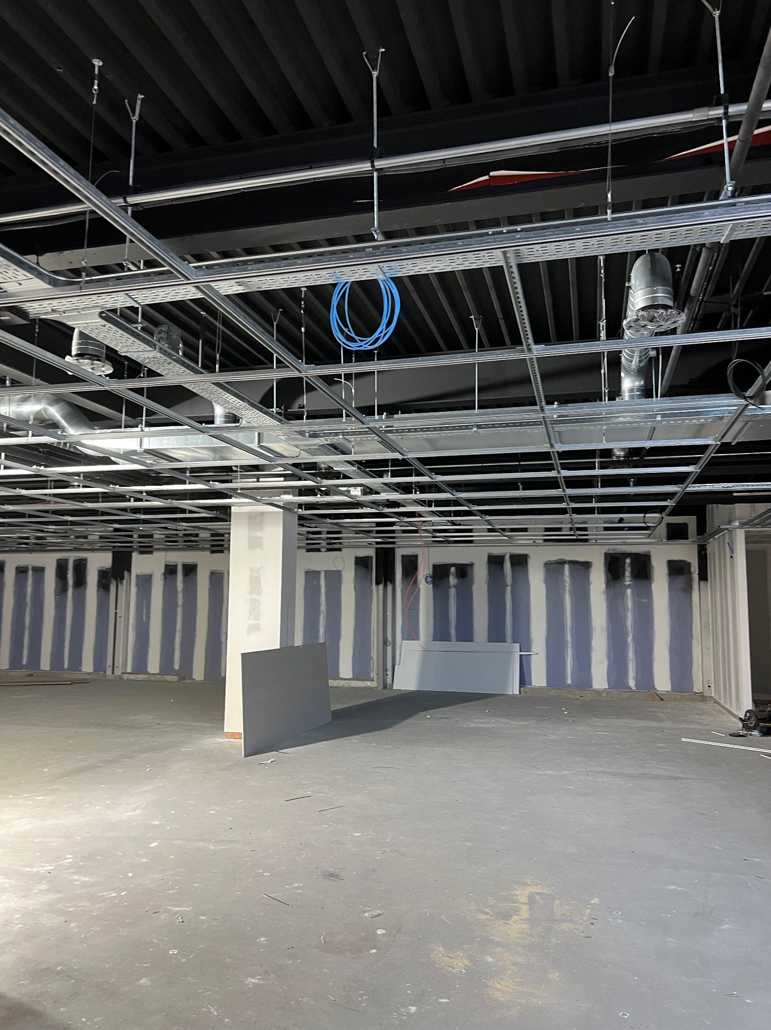 Wnętrze w trakcie prac wykończeniowych: ściany z częściowo zamontowaną konstrukcją nośną, widoczne instalacje pod sufitem, surowy beton na podłodze.