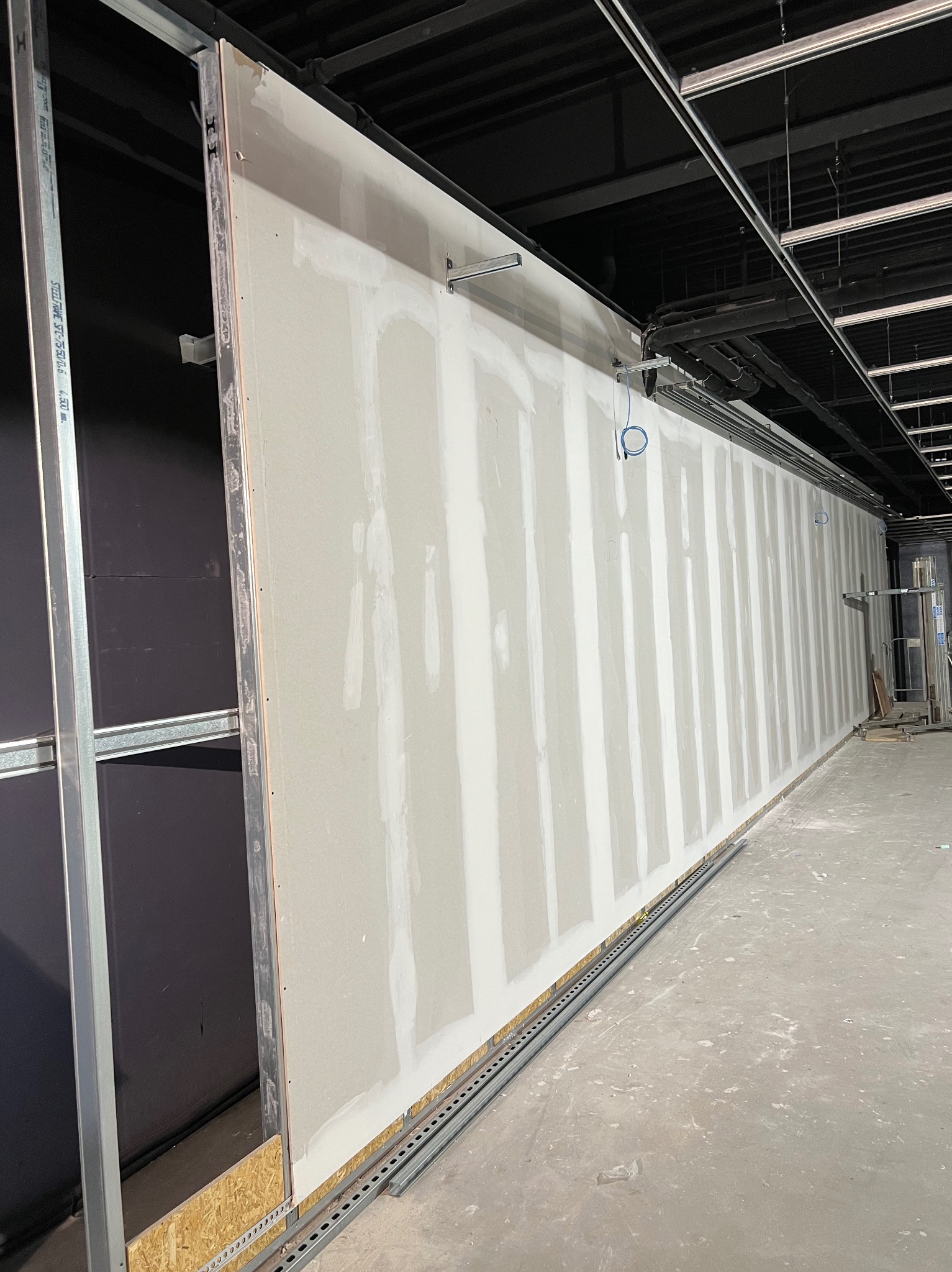 Montaż płyt karton gips na metalowym stelażu w przestrzeni komercyjnej, widoczne niedoskonałości szpachlowania i instalacje podwieszane pod sufitem.