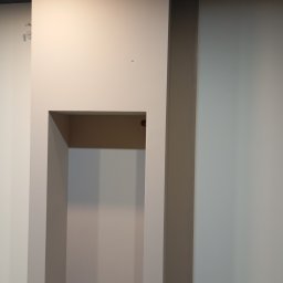 AN-BUD - Wnęka w ścianie pomalowanej na beżowo, z widocznym gniazdkiem elektrycznym u dołu, obok pionowy pas ściany w innym odcieniu beżu, sufit czarny.