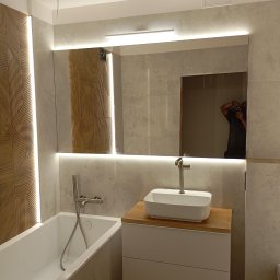 AN-BUD - Nowoczesna łazienka z wanną, umywalką nablatową na drewnianym blacie i dużym lustrem z podświetleniem LED, na ścianach szare płytki imitujące kamień i dekoracyjna drewniana ściana z oświetleniem.