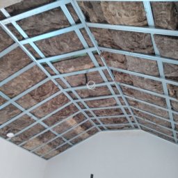 AN-BUD - Konstrukcja sufitu podwieszanego z izolacją z wełny mineralnej, widok od dołu z fragmentem ściany w tle.