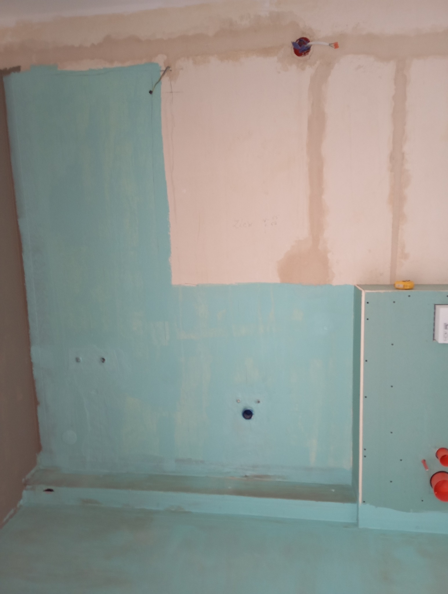 Ściana w trakcie przygotowania do malowania w odcieniach turkusu, widoczne otwory na instalacje, fragment płyty karton-gips z widocznymi wkrętami i pomarańczowymi rurami, surowy stan wykończenia.